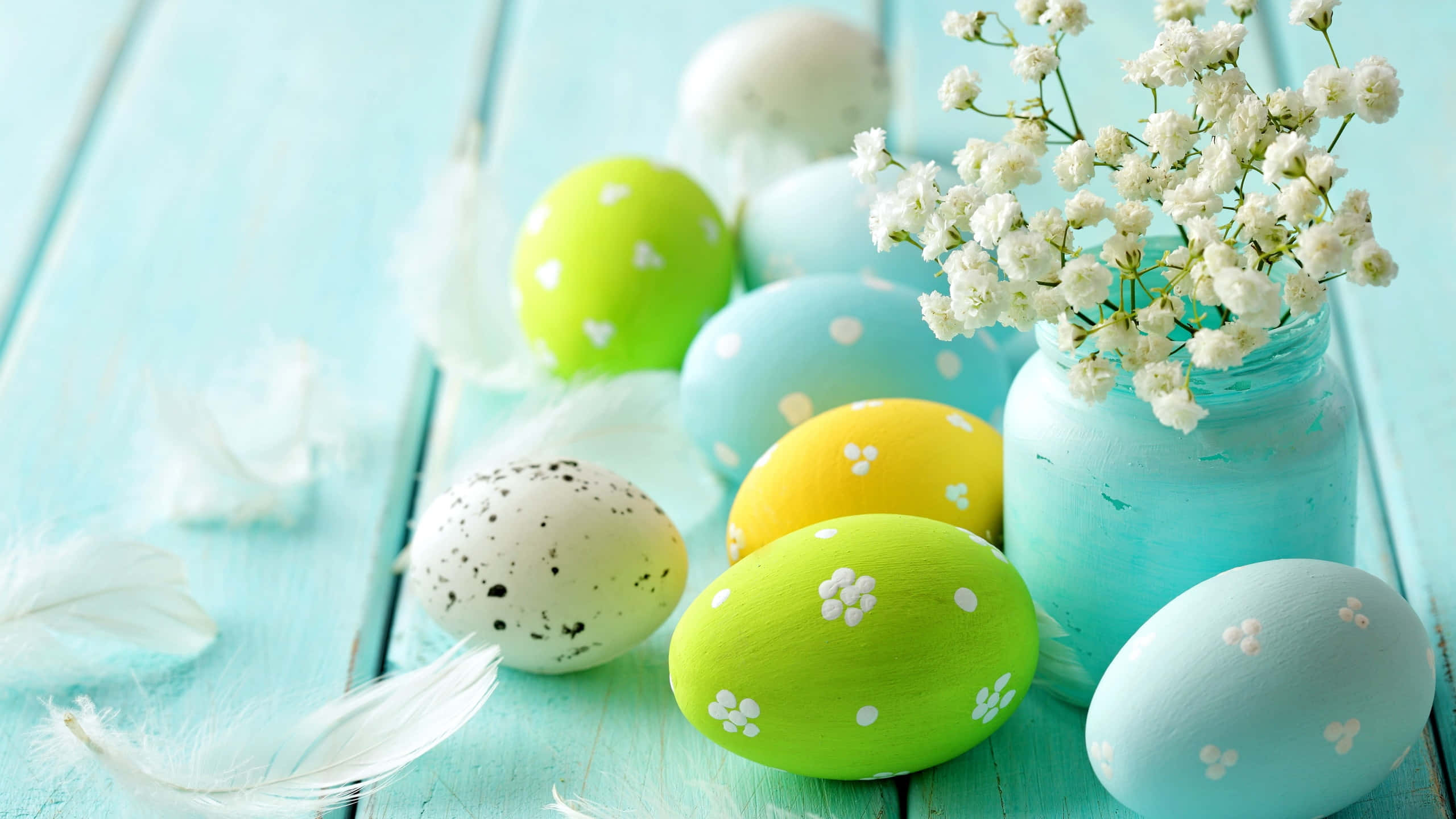 Easter 桌布 壁紙 — 色彩繽紛的手繪復活節蛋和白色滿天星放在一個青綠色的罐子裡，擱在經風化的藍色木質表面上，伴隨著柔軟的白色羽毛。