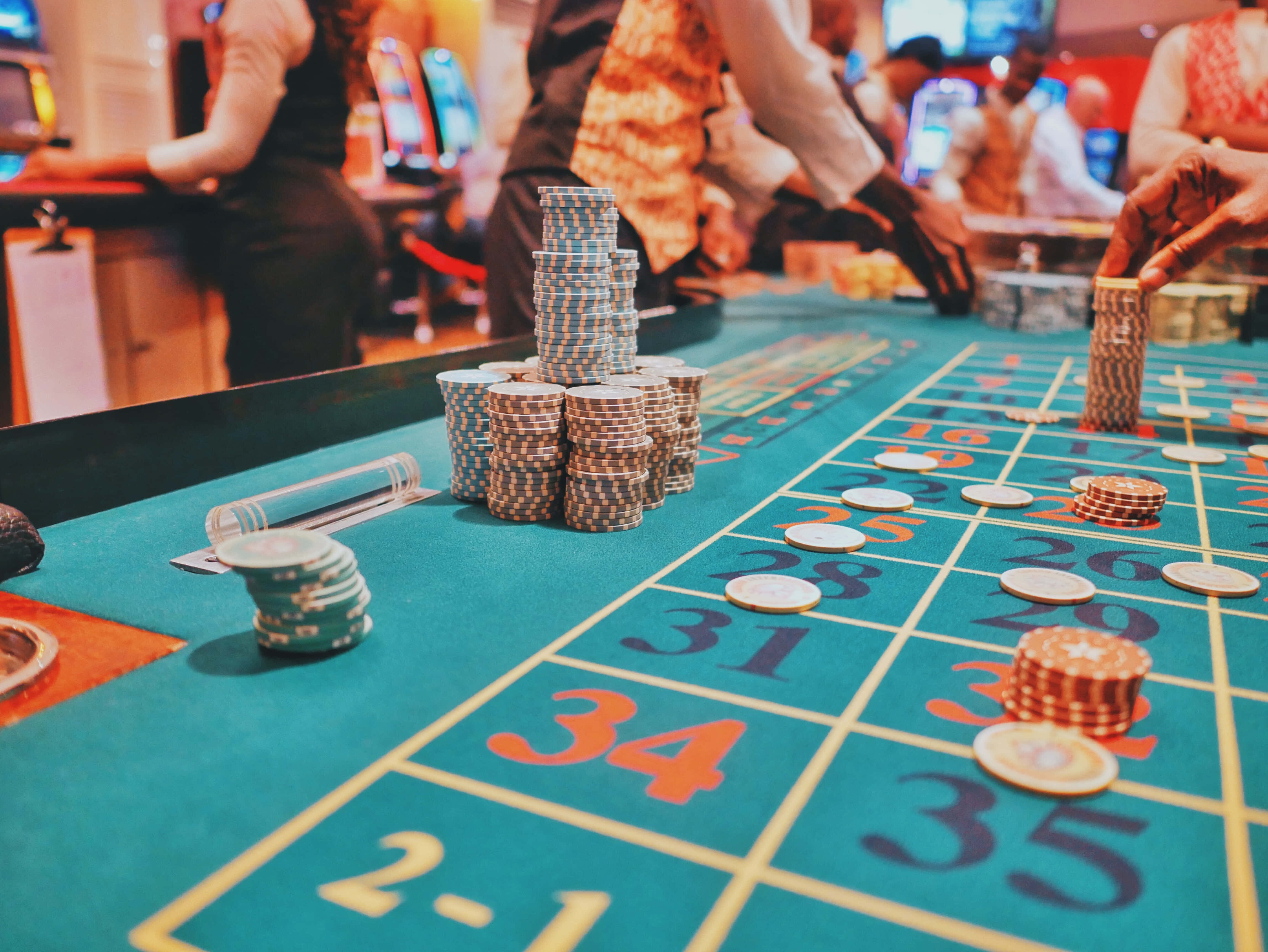 Poker Table 桌布 壁紙 — 色彩繽紛的撲克籌碼堆疊在綠色賭場輪盤桌上的特寫，背景有模糊的玩家。