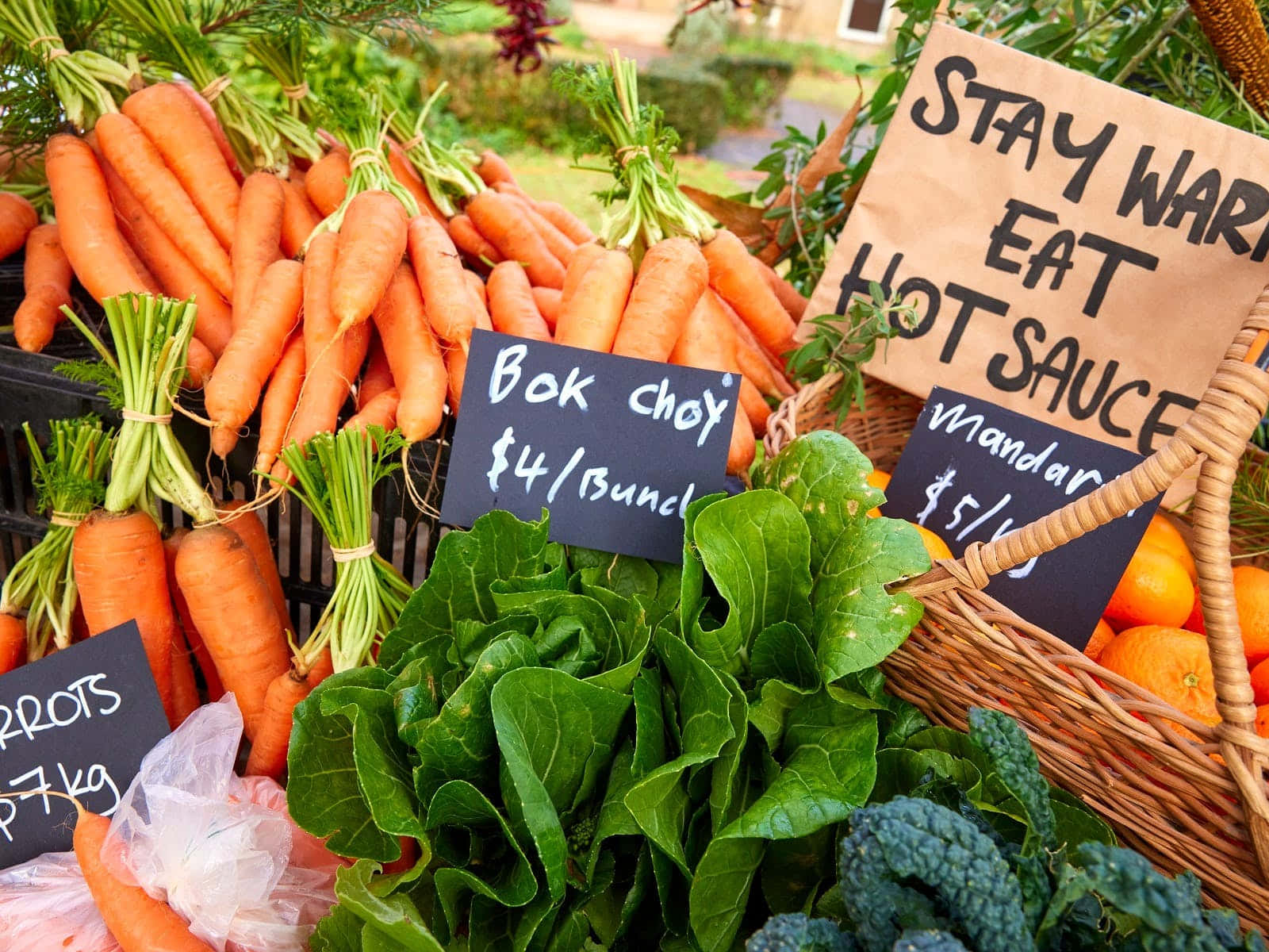 Farmers Market 桌布 壁紙 — 色彩繽紛的新鮮農產品，包括紅蘿蔔、小白菜和柳橙，在戶外農夫市集展示，附有手寫價格標籤。
