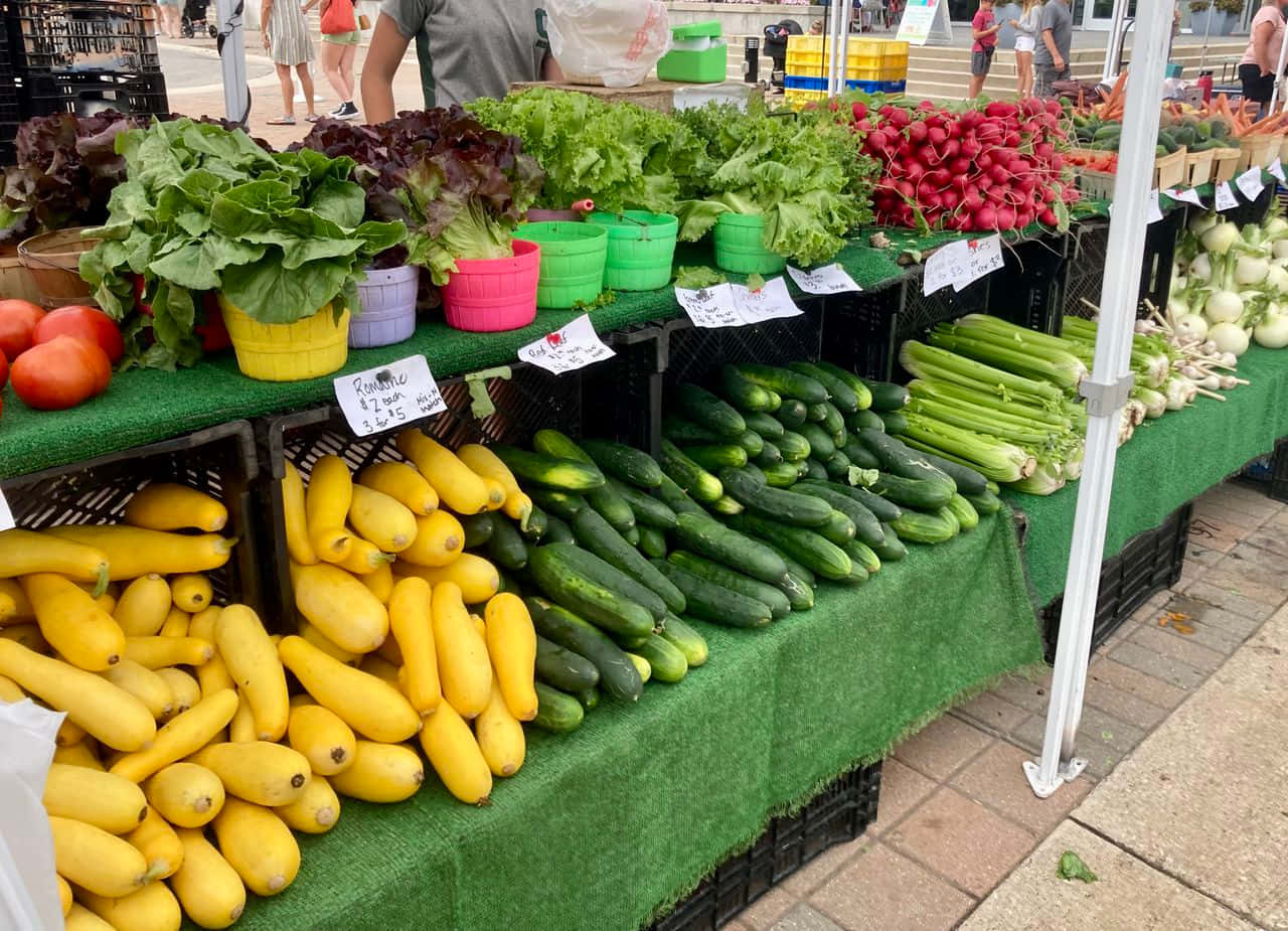 Farmers Market 桌布 壁紙 — 色彩繽紛的新鮮農產品，如黃色南瓜、小黃瓜和蘿蔔，在戶外農夫市場攤位的彩色籃子中展示。