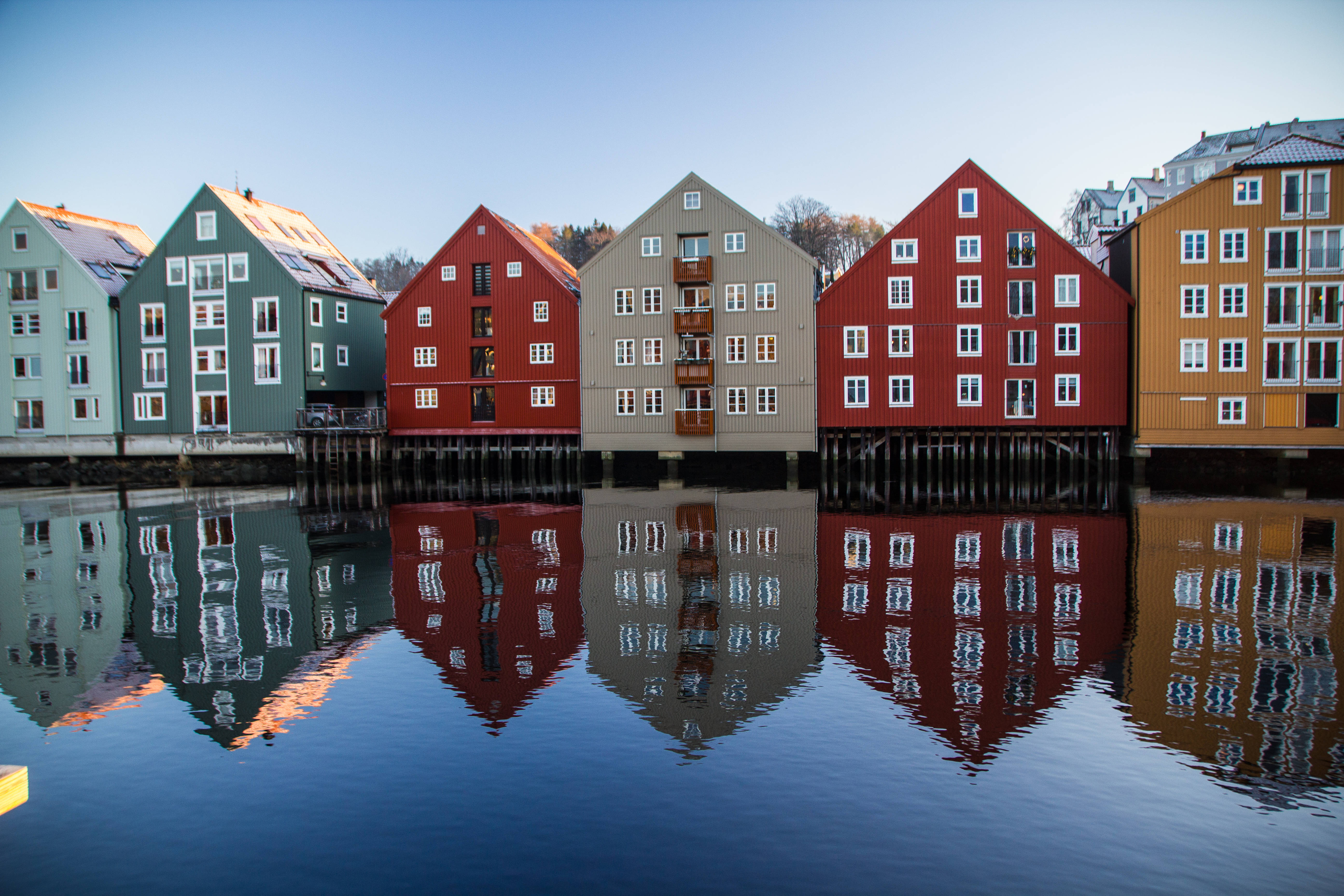Norway 桌布 壁紙 — 色彩繽紛的木屋帶白色窗戶，立於平靜水面的支柱上，在清澈藍天下映照出它們充滿活力的外牆。