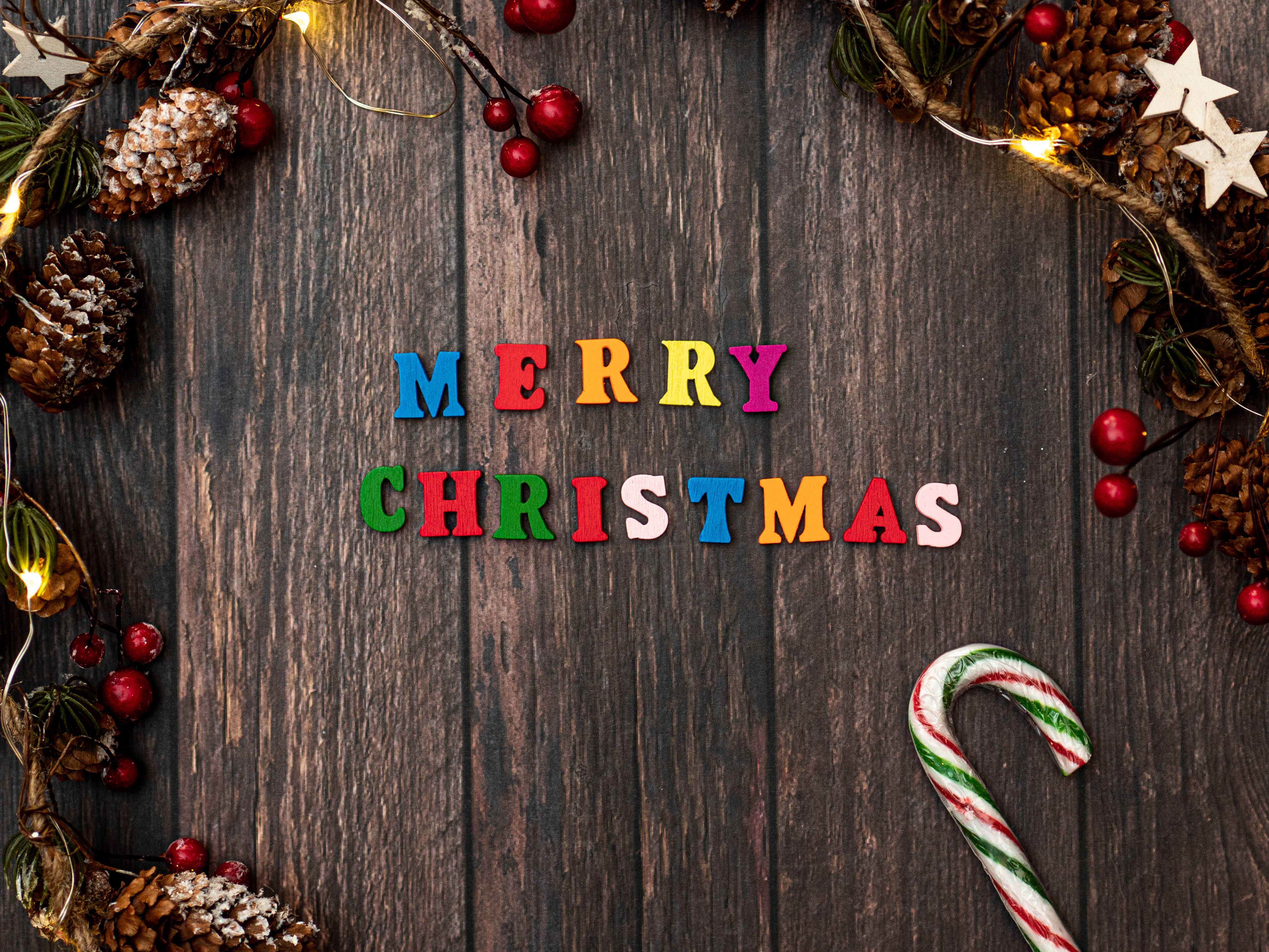 拐杖糖 桌布 壁紙 — 色彩繽紛的木製字母在深色木質表面上拼出「Merry Christmas」，周圍環繞著松果、漿果和一根拐杖糖。