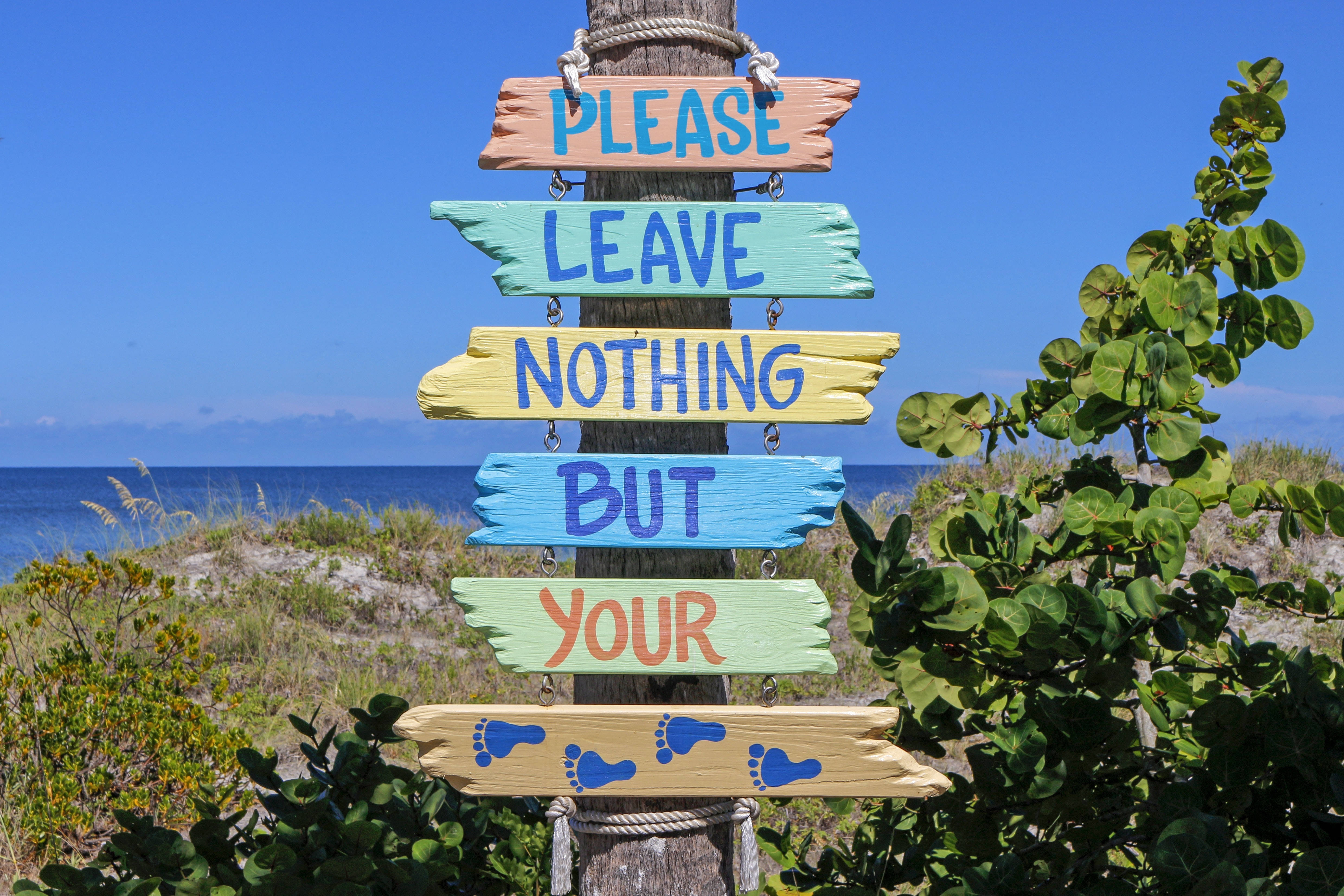 Vacation 桌布 壁紙 — 色彩繽紛的木製標牌上寫著「PLEASE LEAVE NOTHING BUT YOUR FOOTPRINTS」，豎立在沙灘柱上，背景是清澈的藍天和海洋。