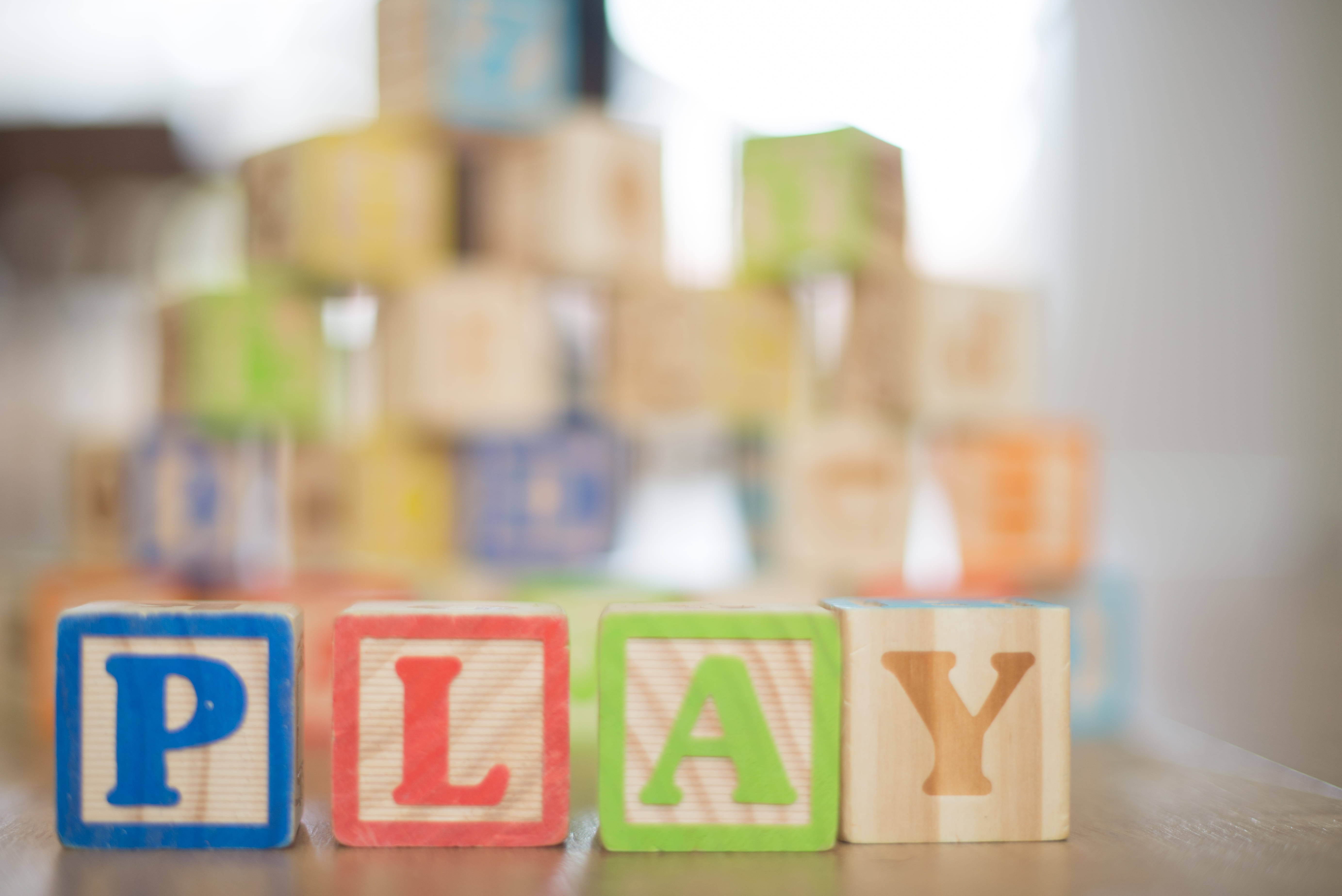 字母A動圖 桌布 壁紙 — 色彩繽紛的木製積木拼出「PLAY」，焦點清晰，背後有模糊的字母積木塔在木質表面上。