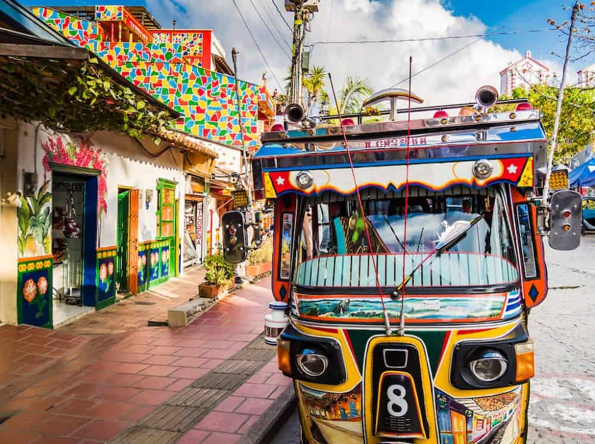 Colombia 桌布 壁紙 — 色彩繽紛的機動三輪車，帶有彩色塗鴉細節，停泊在一條熱鬧的街道上，街道兩旁是藝術裝飾的建築，天空明亮。