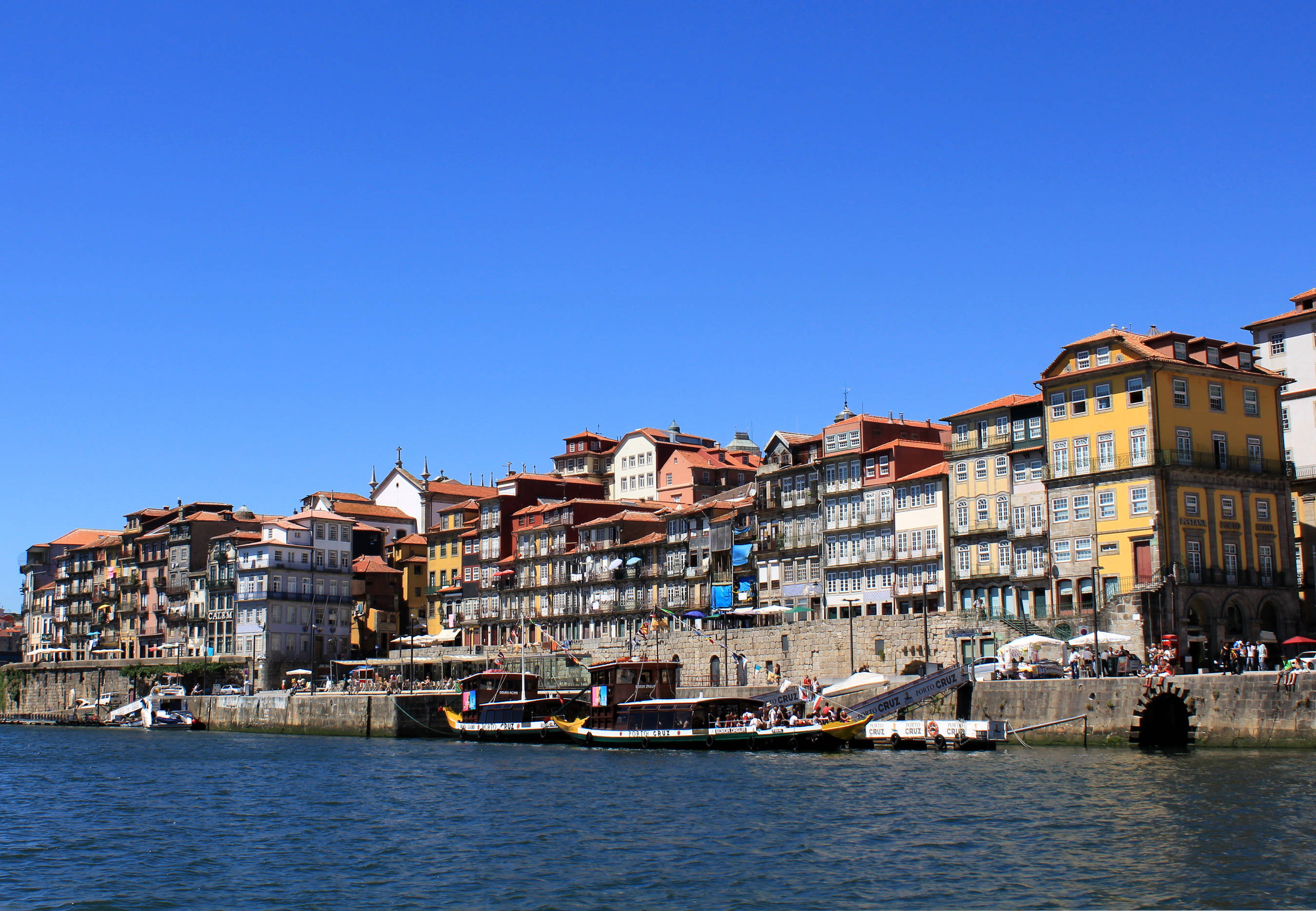Portugal 桌布 壁紙 — 色彩繽紛的歷史建築，擁有陶土屋頂，沿河岸排列，在清澈的藍天下，下方停泊著船隻。