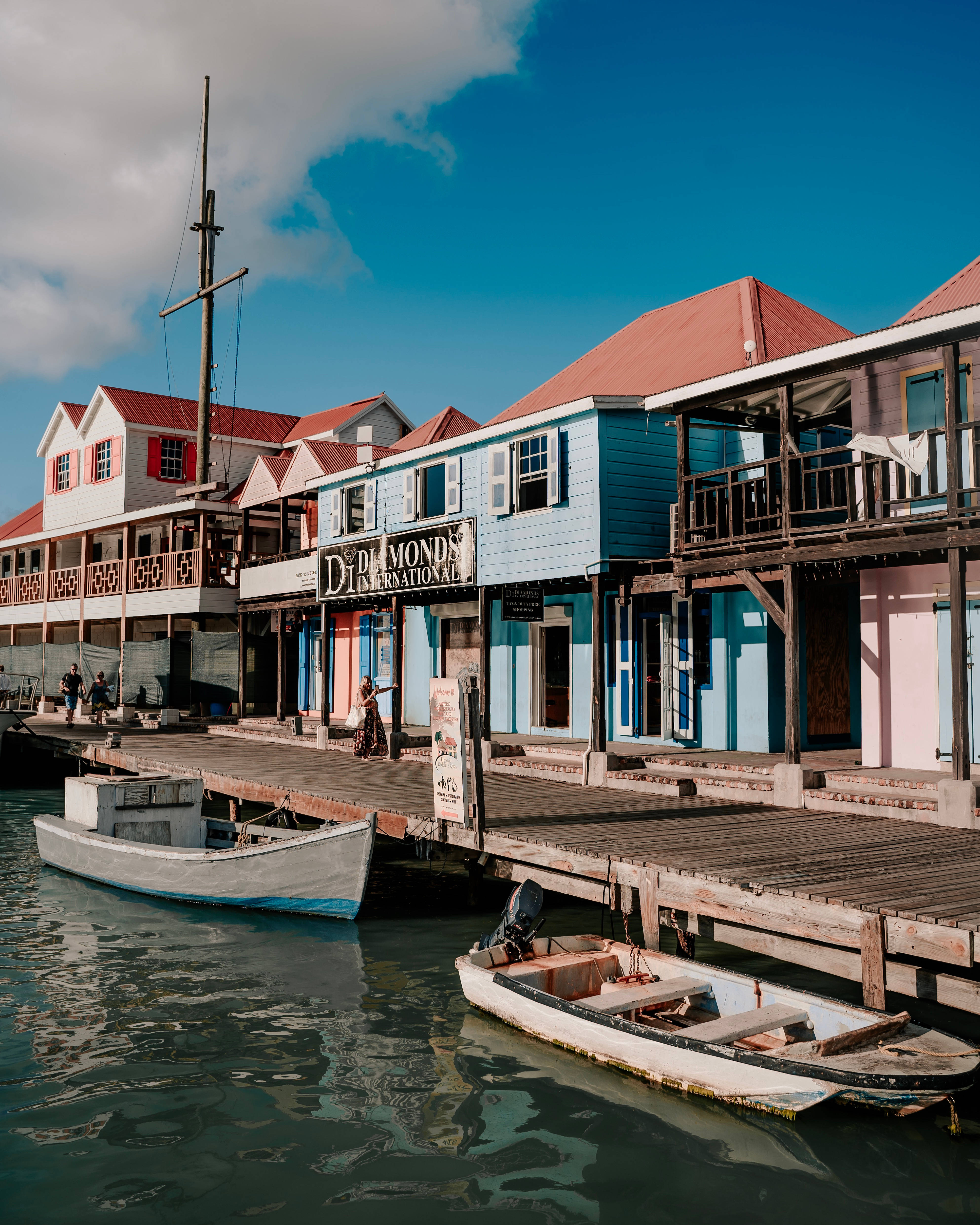 Antigua And Barbuda 桌布 壁紙 — 色彩繽紛的海濱建築物，紅色屋頂，沿著木製碼頭排列，小船在平靜的碧綠海水中停泊，天空湛藍明亮。
