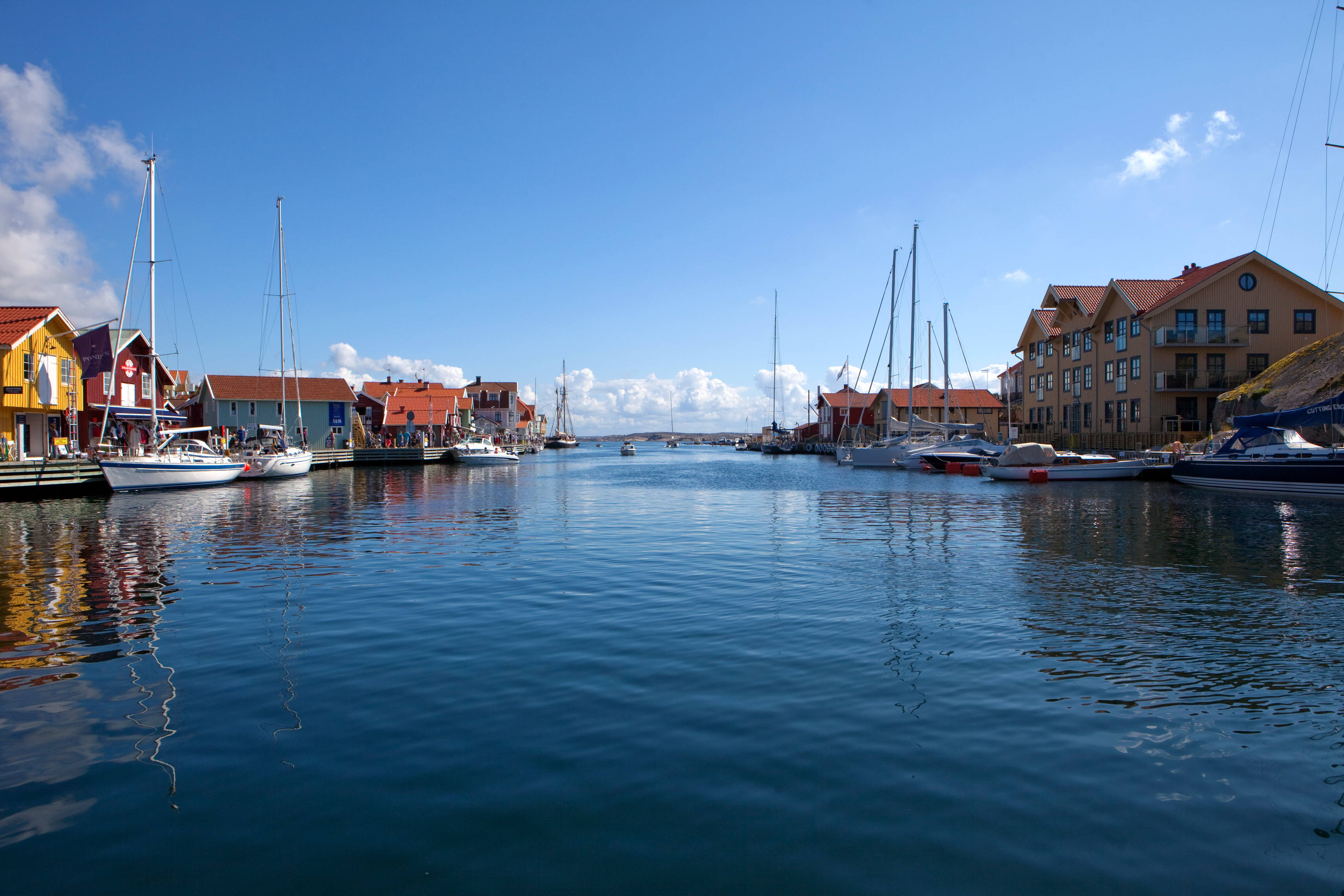 Sweden 桌布 壁紙 — 色彩繽紛的海濱房屋排列在平靜的港口旁，帆船點綴其間，天空湛藍，白雲朵朵。
