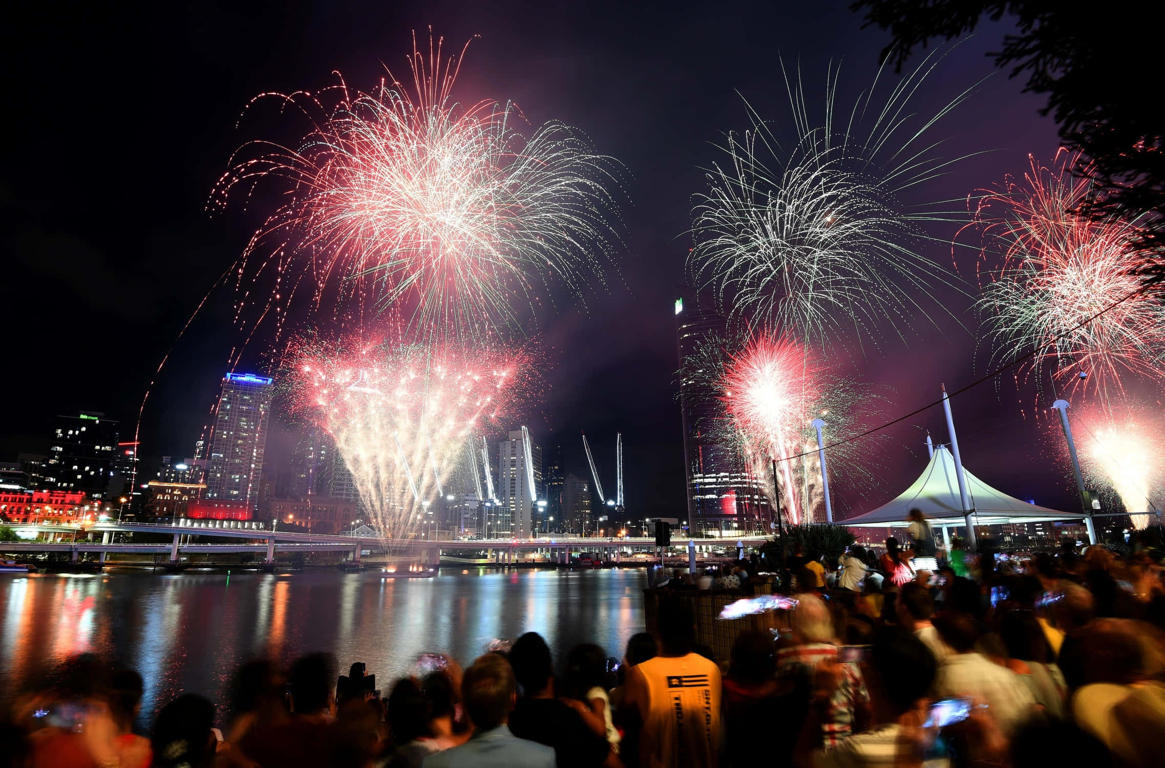 New Years Eve 桌布 壁紙 — 色彩繽紛的煙火在倒映在水中的城市天際線上爆炸，人群在夜晚從岸邊觀看。