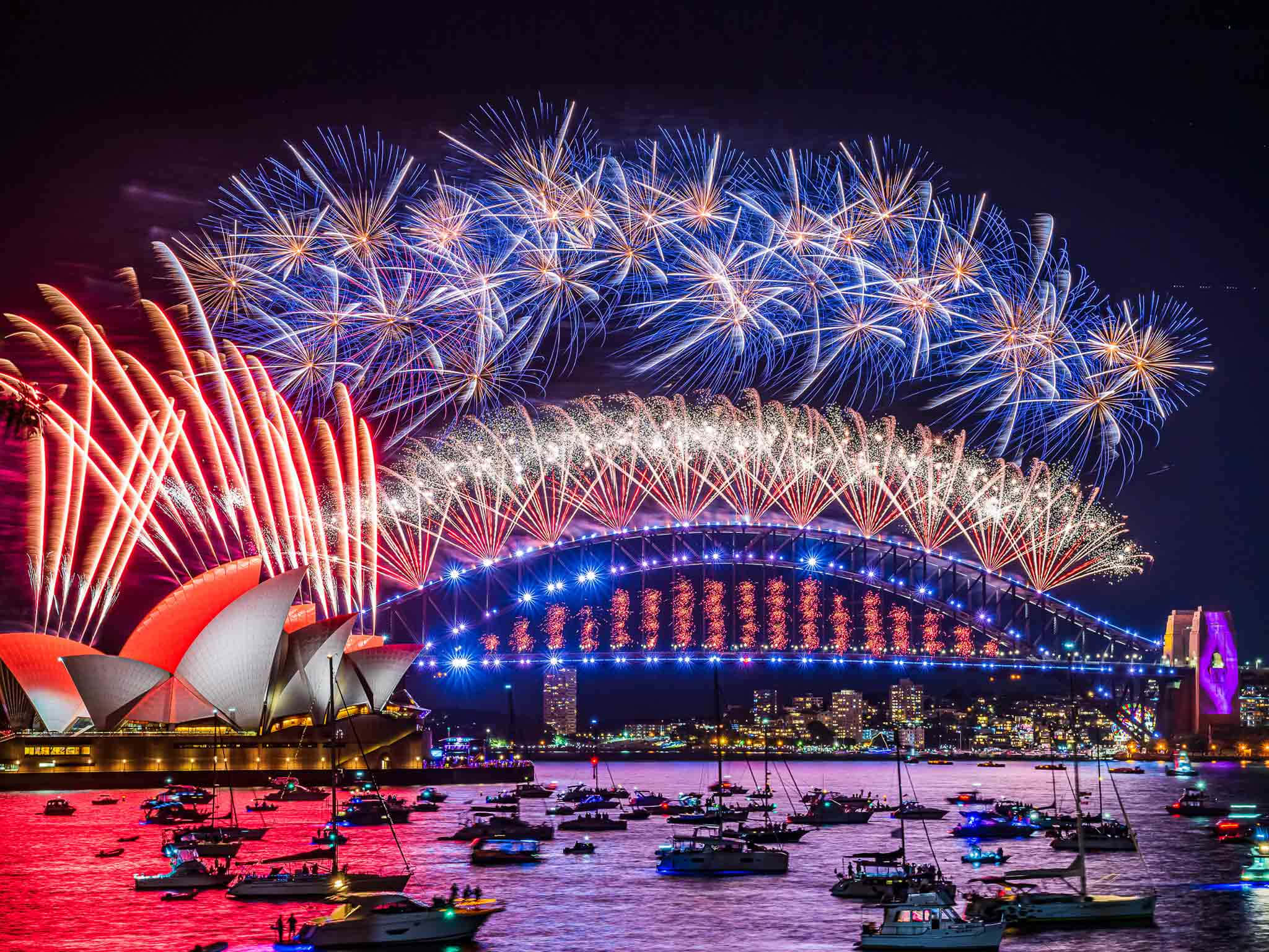 新年 桌布 壁紙 — 色彩繽紛的煙火在夜間於雪梨港上空綻放，照亮了歌劇院和港灣大橋，下方有船隻。