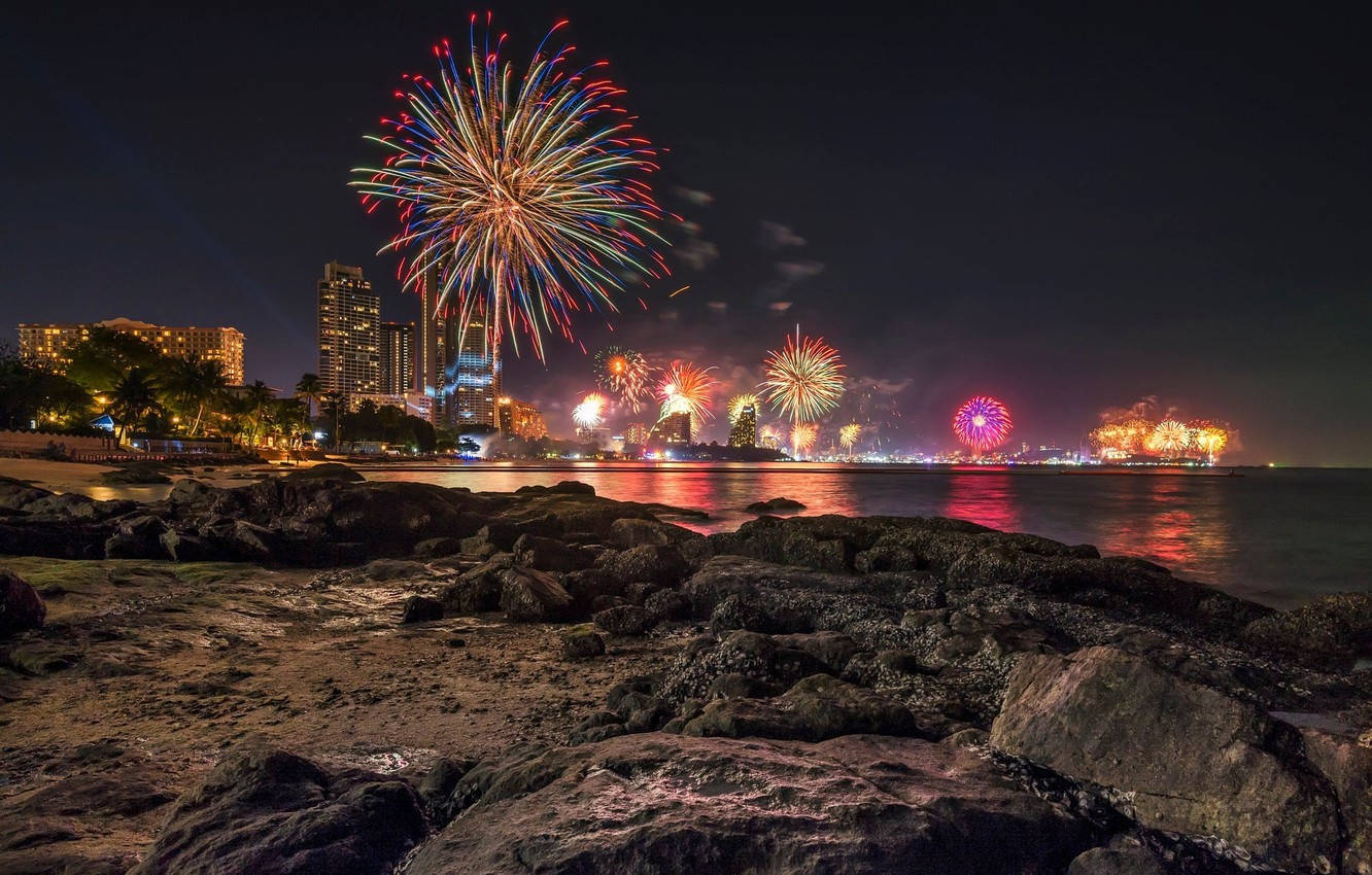 Fireworks 桌布 壁紙 — 色彩繽紛的煙火在夜間沿海城市的天際線上綻放，倒映在水面上，前景是岩石。
