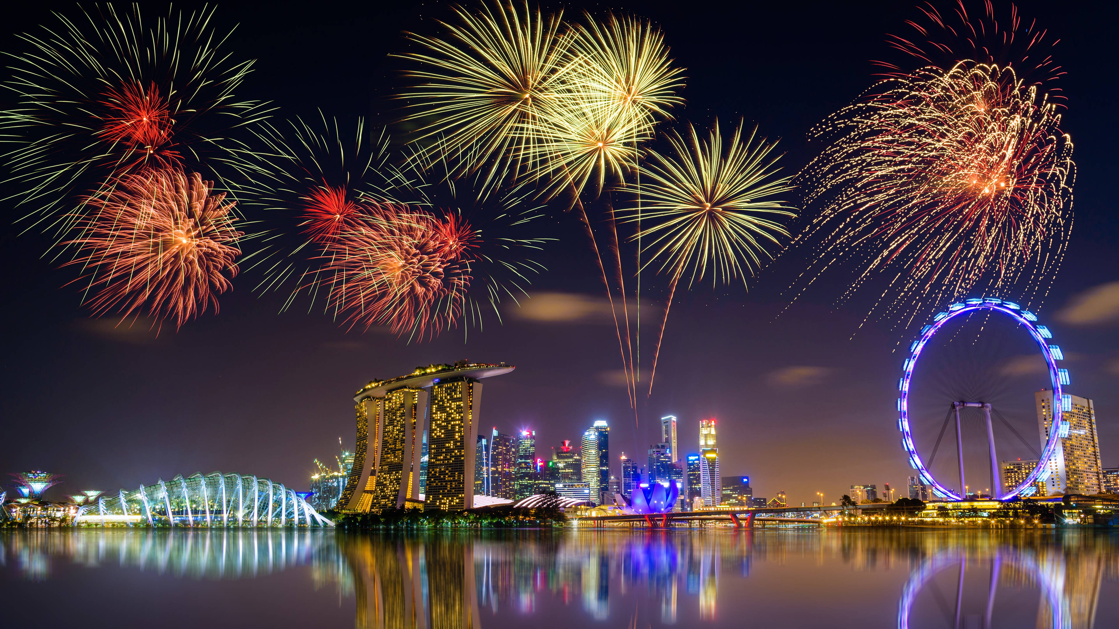 Singapore 桌布 壁紙 — 色彩繽紛的煙火在新加坡璀璨的天際線上綻放，濱海灣金沙和新加坡摩天觀景輪倒映在平靜的水面上。
