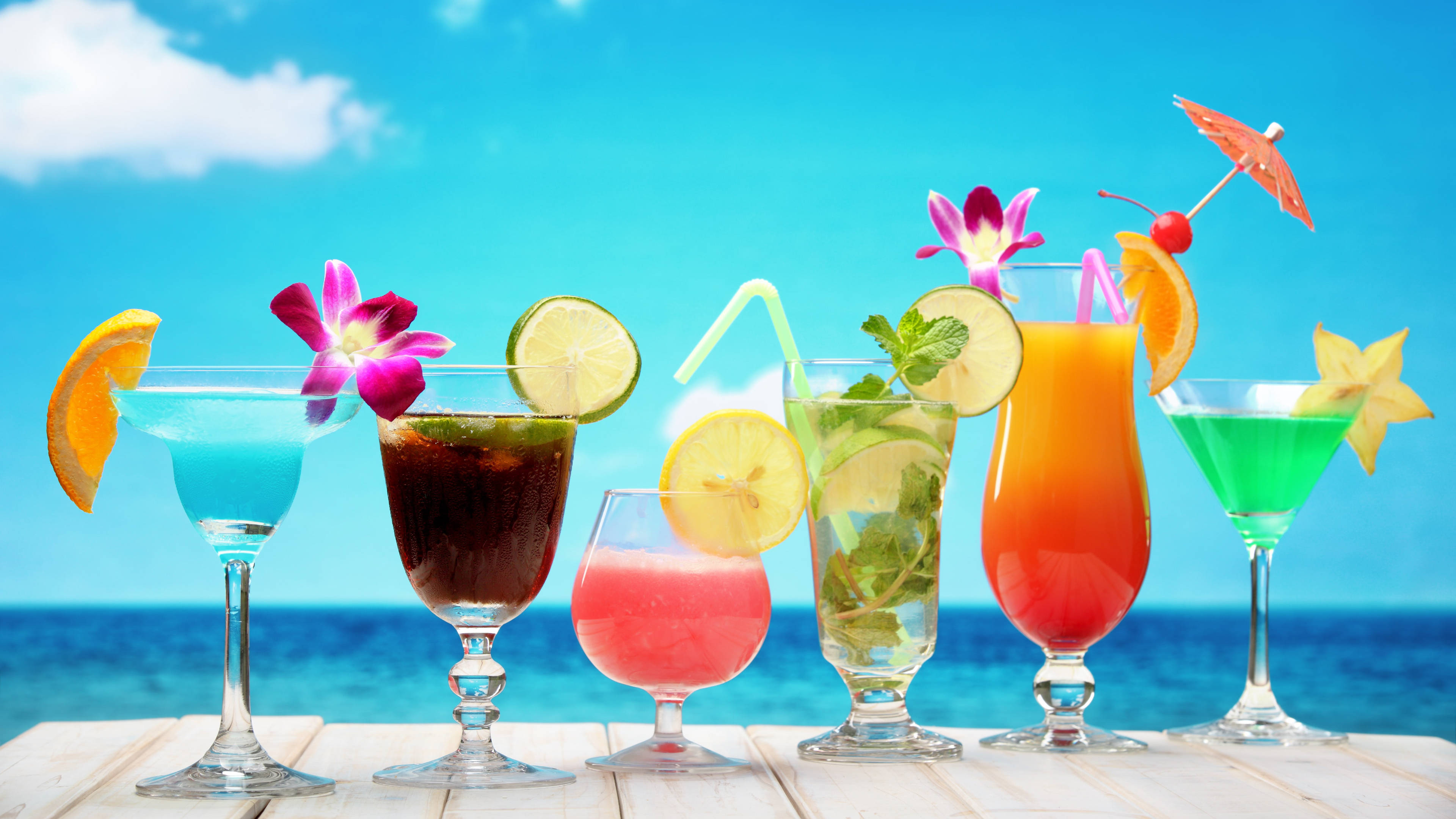 Tropical Drink 桌布 壁紙 — 色彩繽紛的熱帶雞尾酒配上水果裝飾和鮮花，放在木桌上，背景是明亮的藍色海洋和天空。