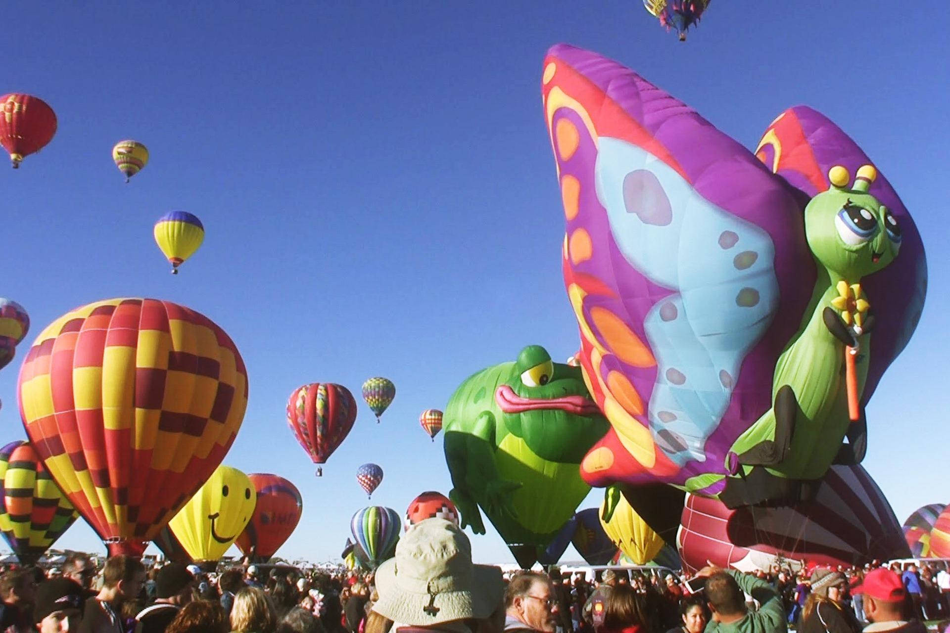 Balloon 桌布 壁紙 — 色彩繽紛的熱氣球，形狀像蝴蝶、青蛙和格子圖案，在晴朗的藍天下飄浮在歡呼的人群上方。
