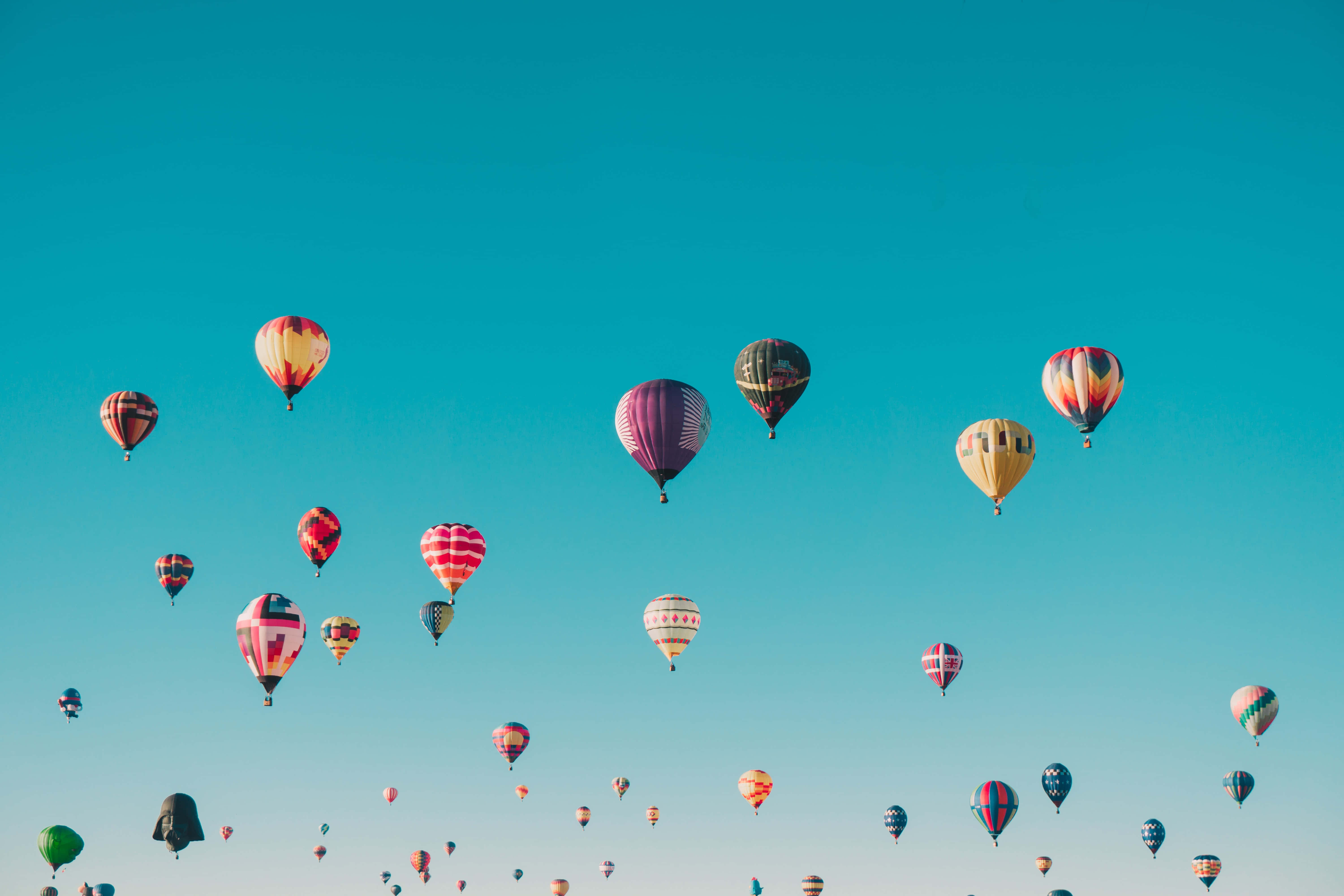 Hot Air Balloon 桌布 壁紙 — 色彩繽紛的熱氣球以各種圖案在清澈的藍綠色天空中飄浮，呈現節日般的空中表演。
