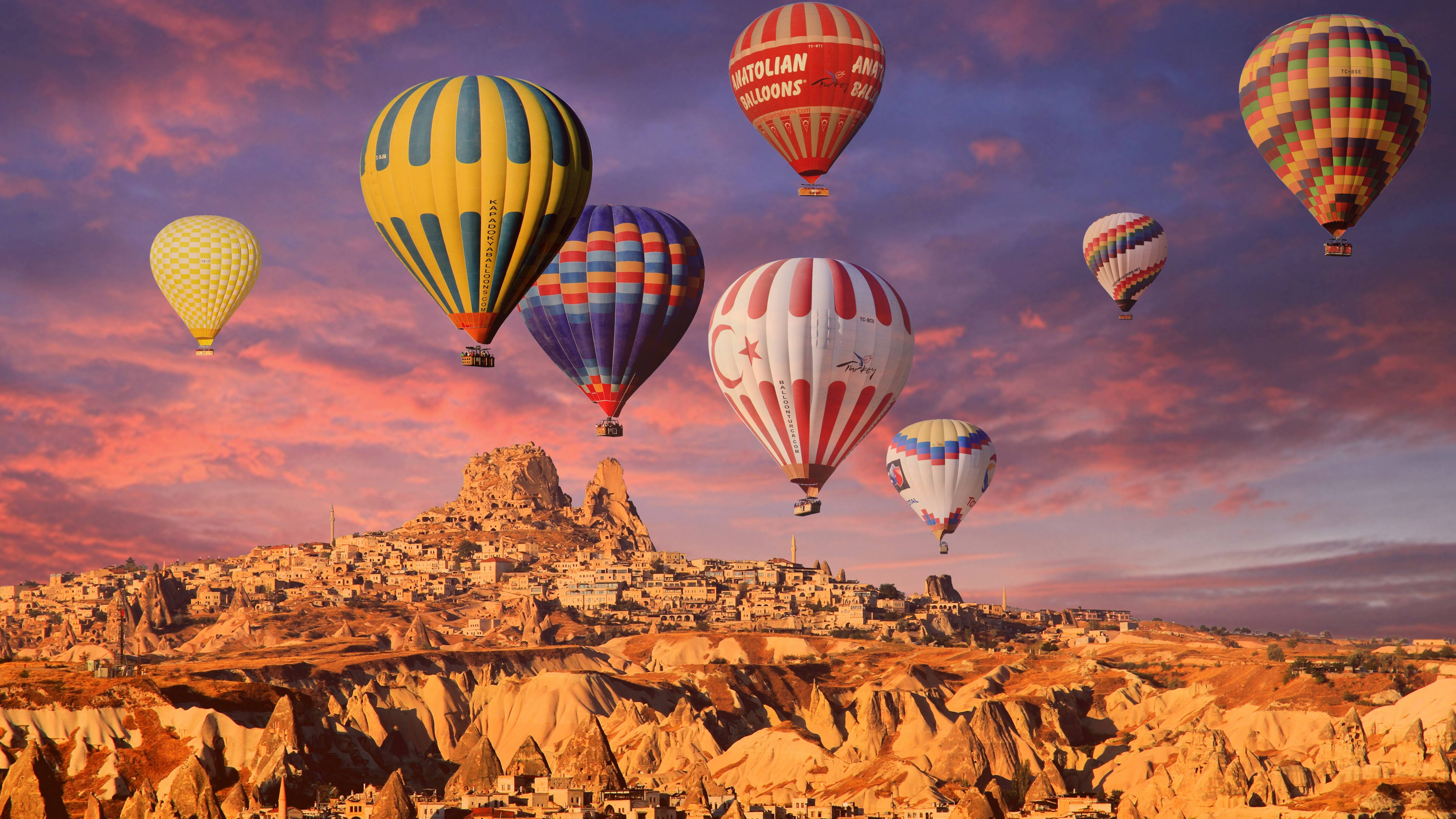 Cappadocia 桌布 壁紙 — 色彩繽紛的熱氣球在日落時分飄過崎嶇的山谷，位於歷史悠久的懸崖小鎮之上。