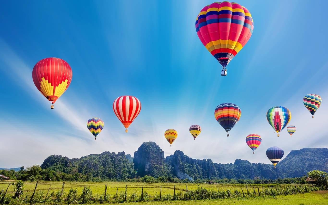 Hot Air Balloon 桌布 壁紙 — 色彩繽紛的熱氣球在晴朗的藍天上翱翔，下方是鬱鬱蔥蔥的綠色田野和崎嶇的石灰岩山脈。