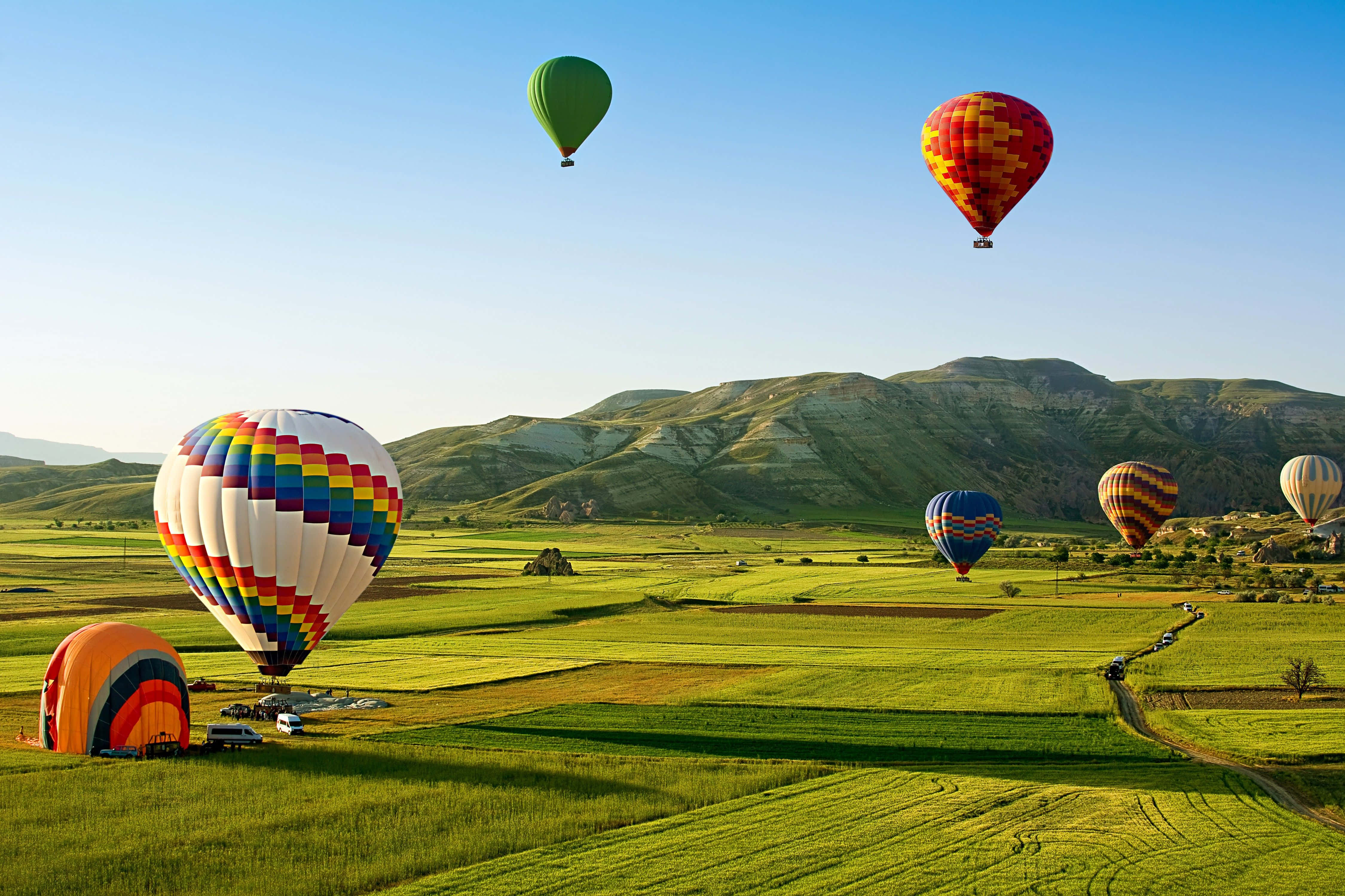Hot Air Balloon 桌布 壁紙 — 色彩繽紛的熱氣球在晴朗的藍天下，飄過陽光普照的綠色田野和起伏的丘陵。