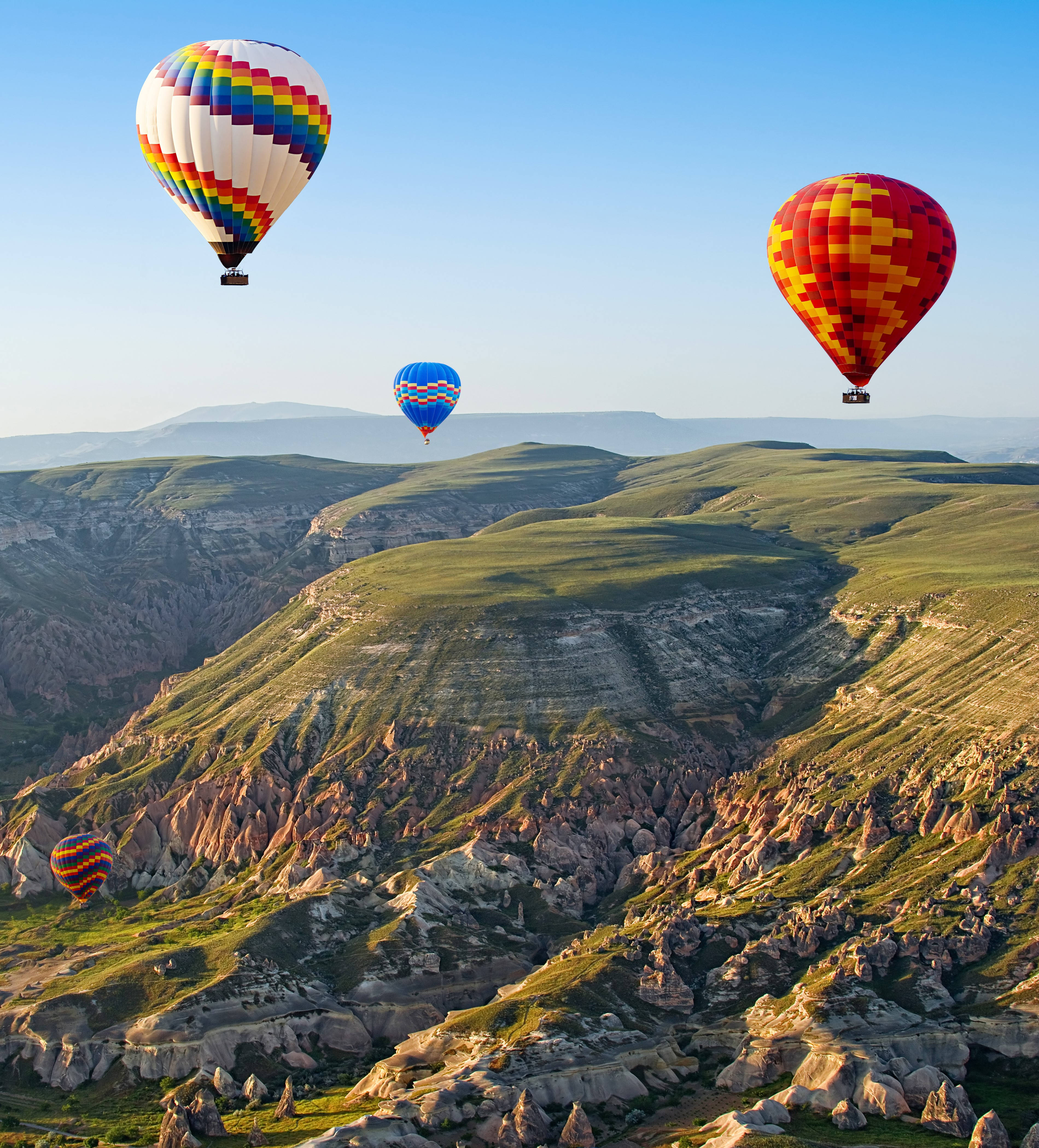 Cappadocia 桌布 壁紙 — 色彩繽紛的熱氣球在晴朗的藍天下飄過崎嶇的綠色山丘和岩石地貌。