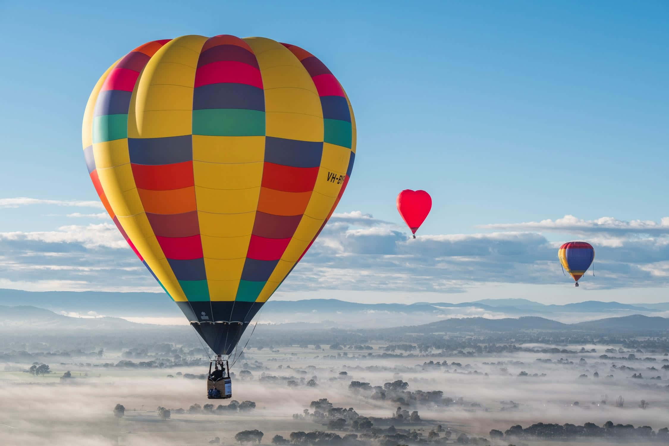 Hot Air Balloon 桌布 壁紙 — 色彩繽紛的熱氣球在晴朗藍天下，飛越霧氣瀰漫的山谷和起伏的丘陵。