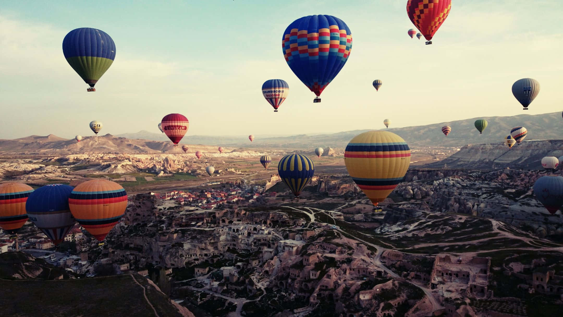 Hot Air Balloon 桌布 壁紙 — 色彩繽紛的熱氣球在柔和粉彩天空下，於黃金時刻飄過崎嶇的山谷小鎮。