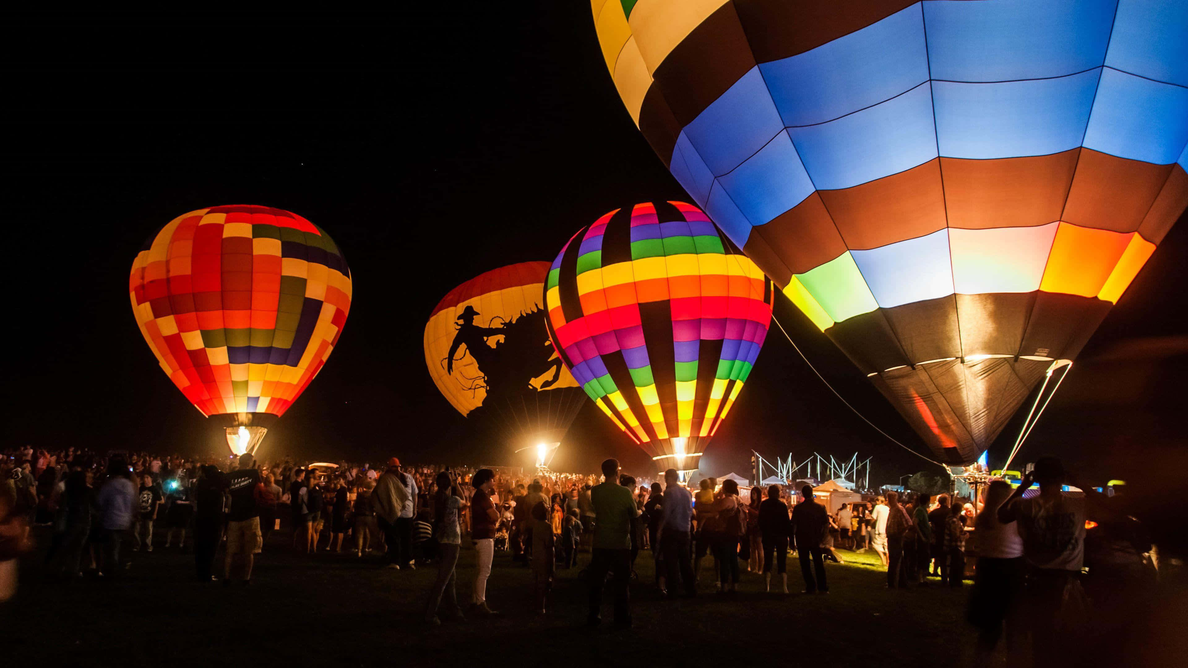 Hot Air Balloon 桌布 壁紙 — 色彩繽紛的熱氣球在漆黑的夜空中閃耀著彩虹色調，人群在下方聚集。