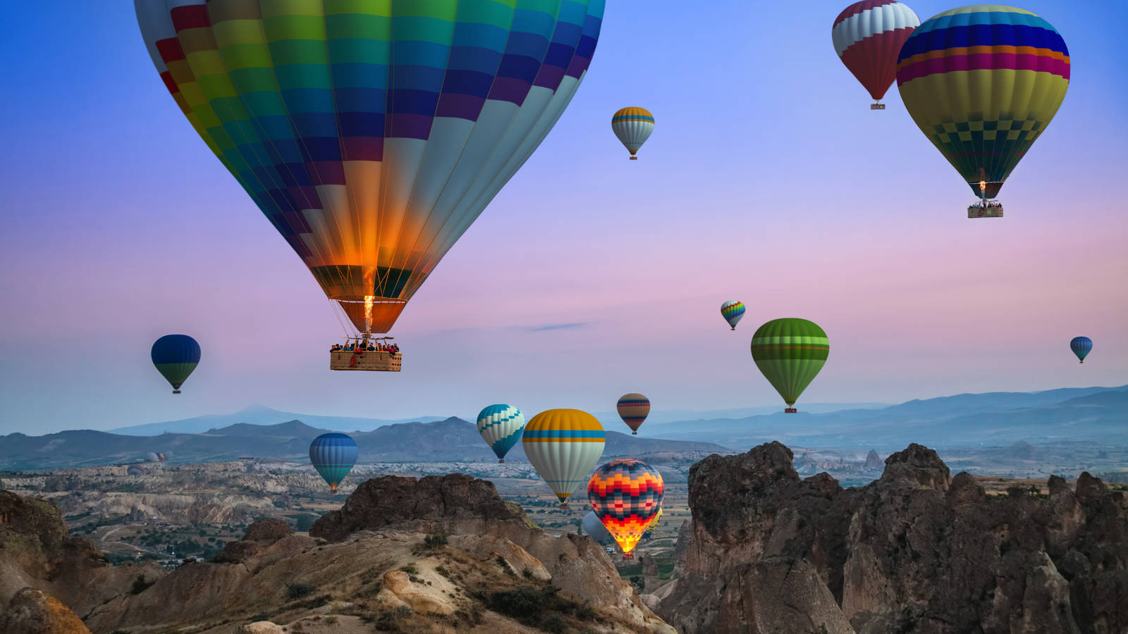 Cappadocia 桌布 壁紙 — 色彩繽紛的熱氣球在黃昏時分飄過崎嶇的火山岩層，天空呈現柔和的粉紅色和藍色。