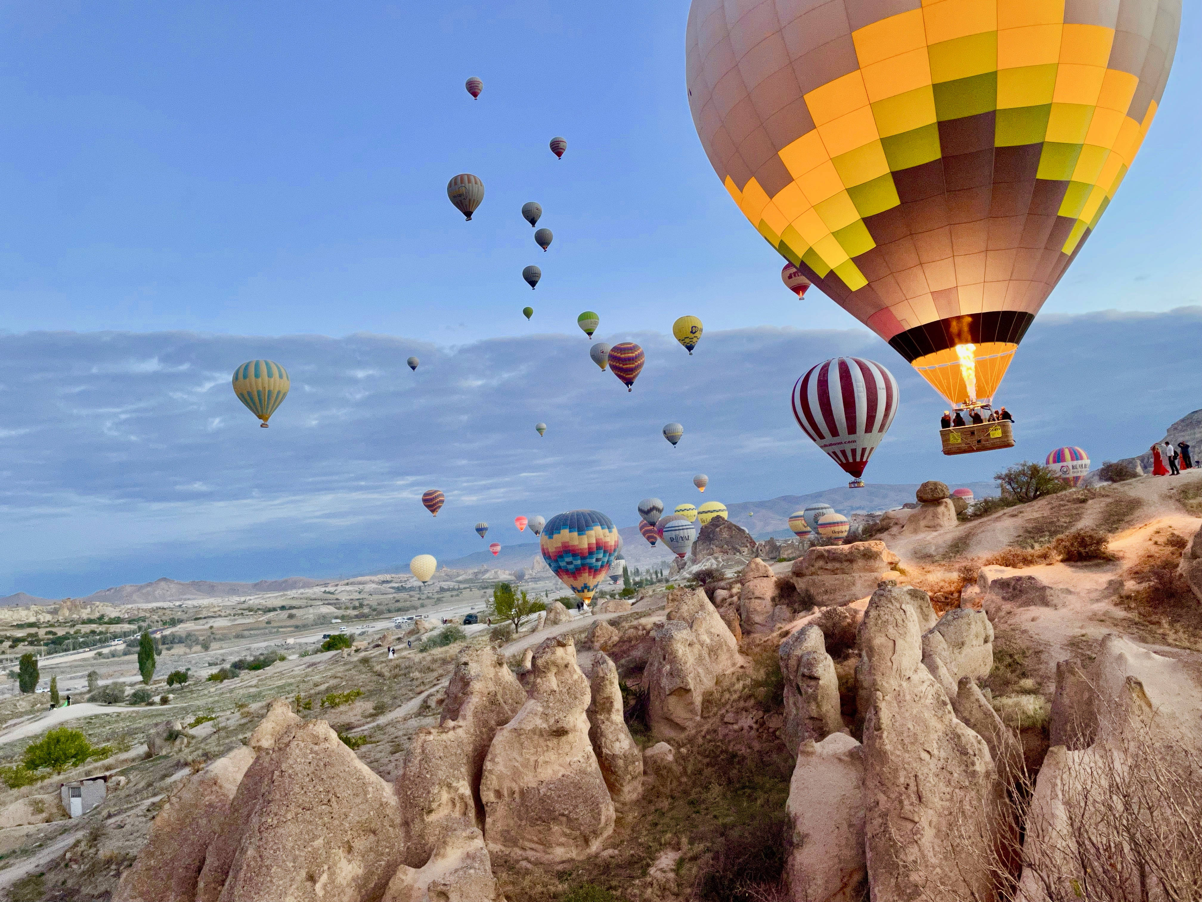 Cappadocia 桌布 壁紙 — 色彩繽紛的熱氣球在黎明時分柔和的藍天下飄過崎嶇的岩石地貌。