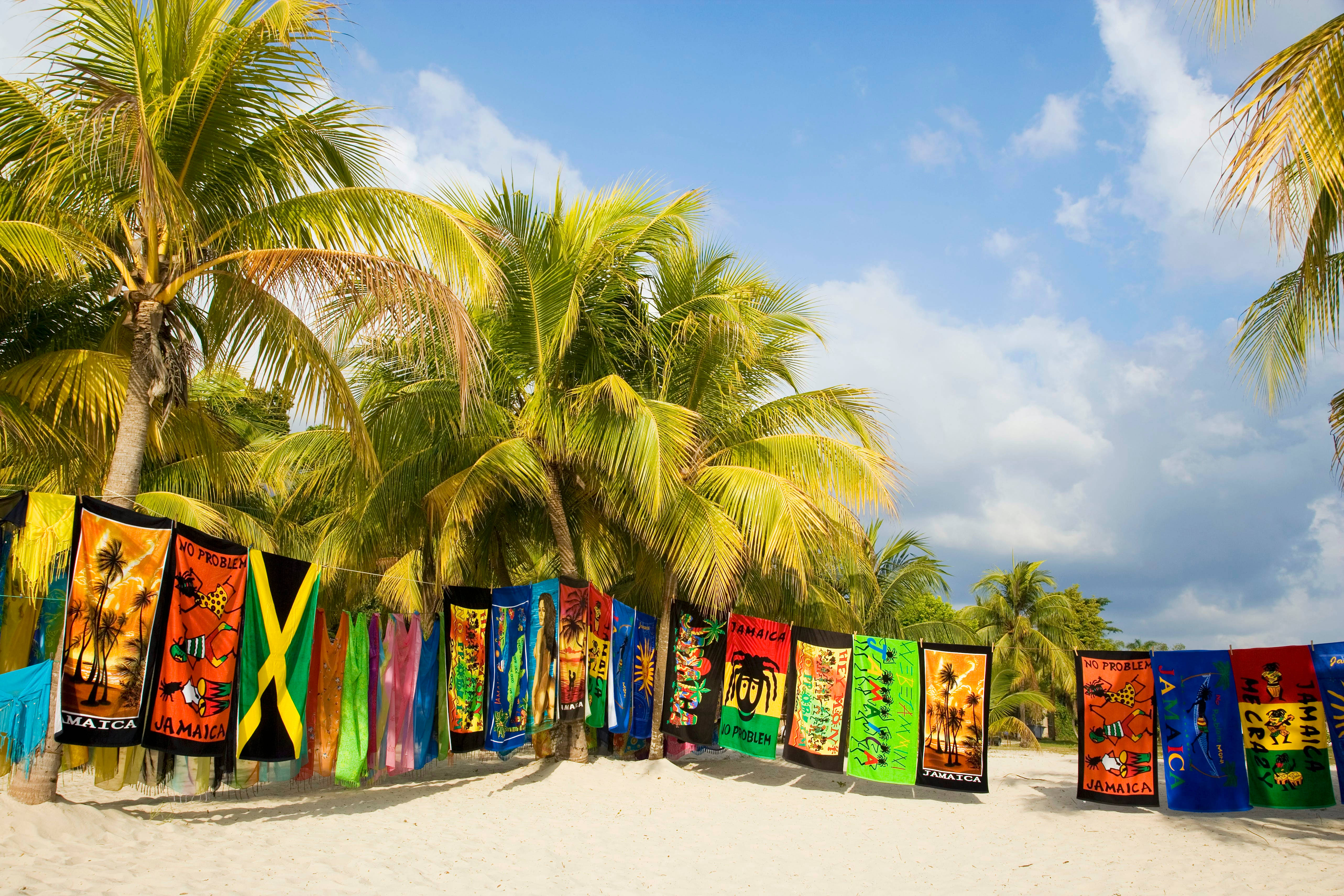 Jamaica 桌布 壁紙 — 色彩繽紛的牙買加沙灘毛巾掛在棕櫚樹之間，位於陽光明媚的熱帶海岸，藍天白雲。