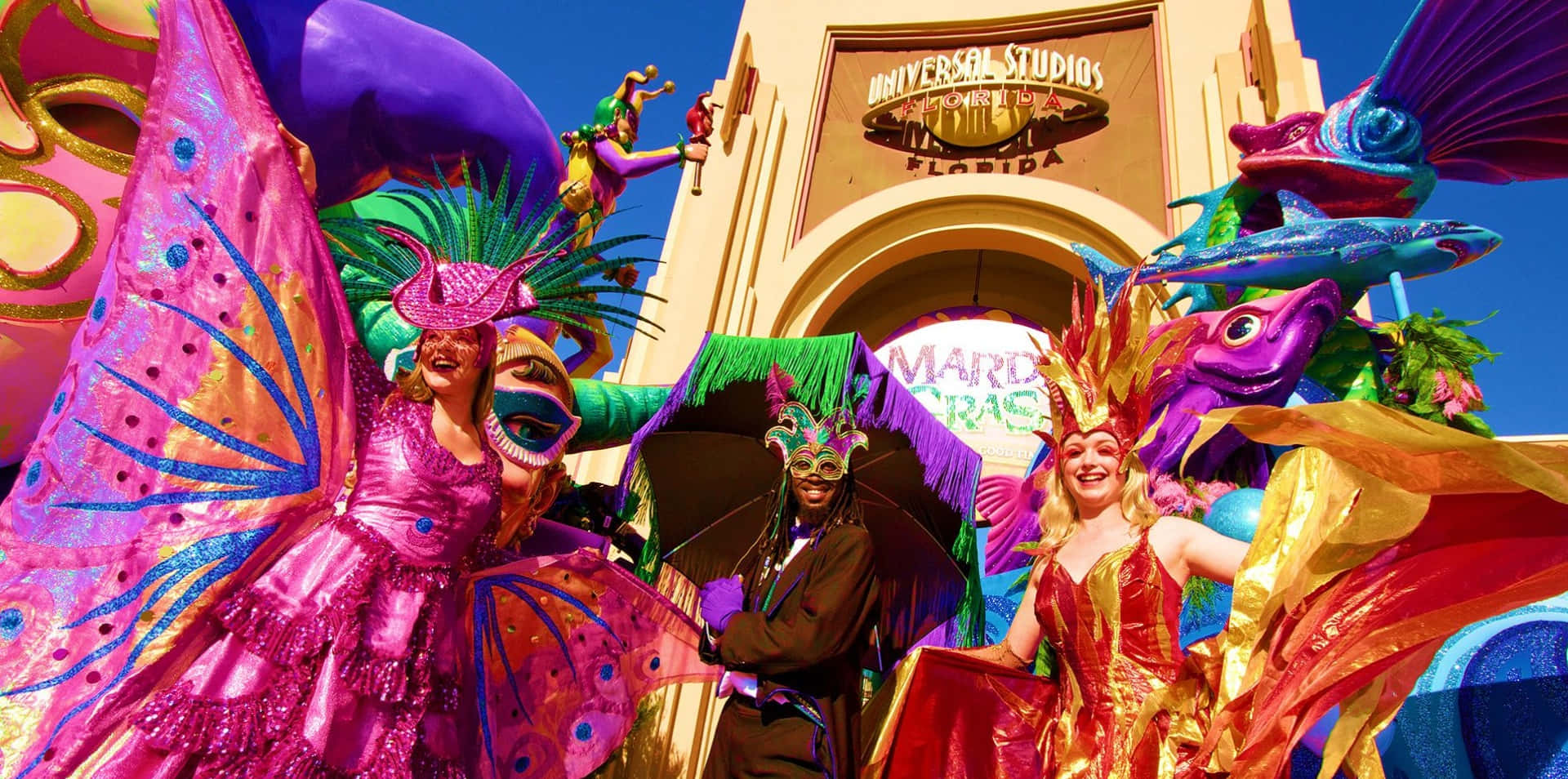 Mardi Gras 桌布 壁紙 — 色彩繽紛的狂歡節表演者身穿閃亮服飾，在晴朗藍天下於佛羅里達環球影城入口前遊行。