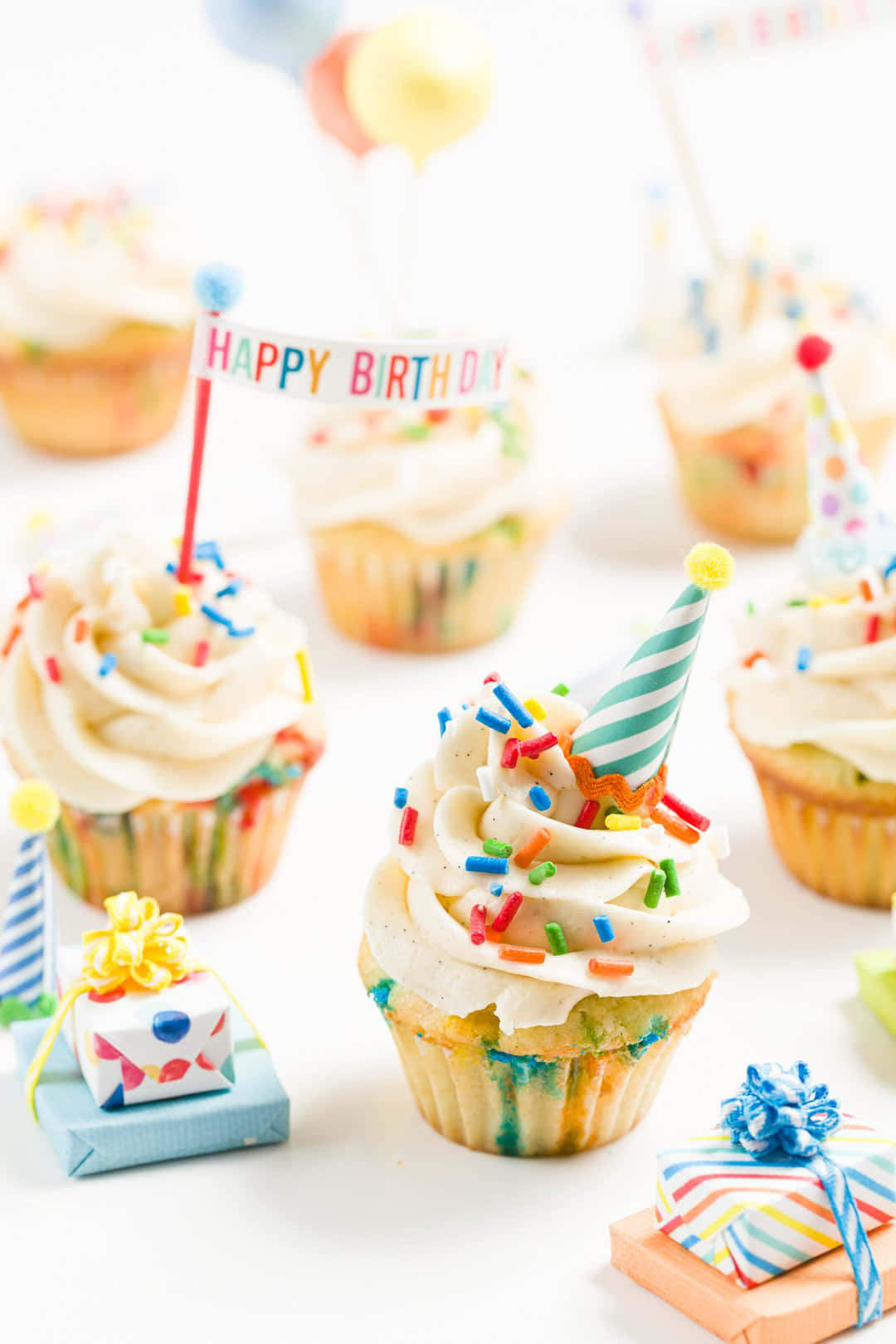 Cupcake 桌布 壁紙 — 色彩繽紛的生日紙杯蛋糕，配有糖霜、派對帽和禮物盒，排列在明亮的白色表面上。