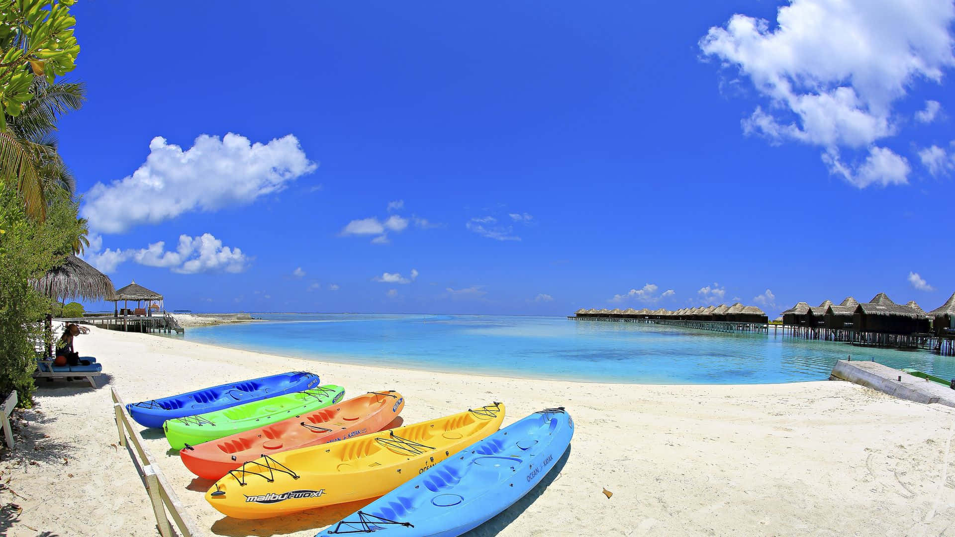Tropical Beach 桌布 壁紙 — 色彩繽紛的皮划艇排列在純淨的白色沙灘上，天空湛藍，白雲朵朵，遠處還有水上平房。