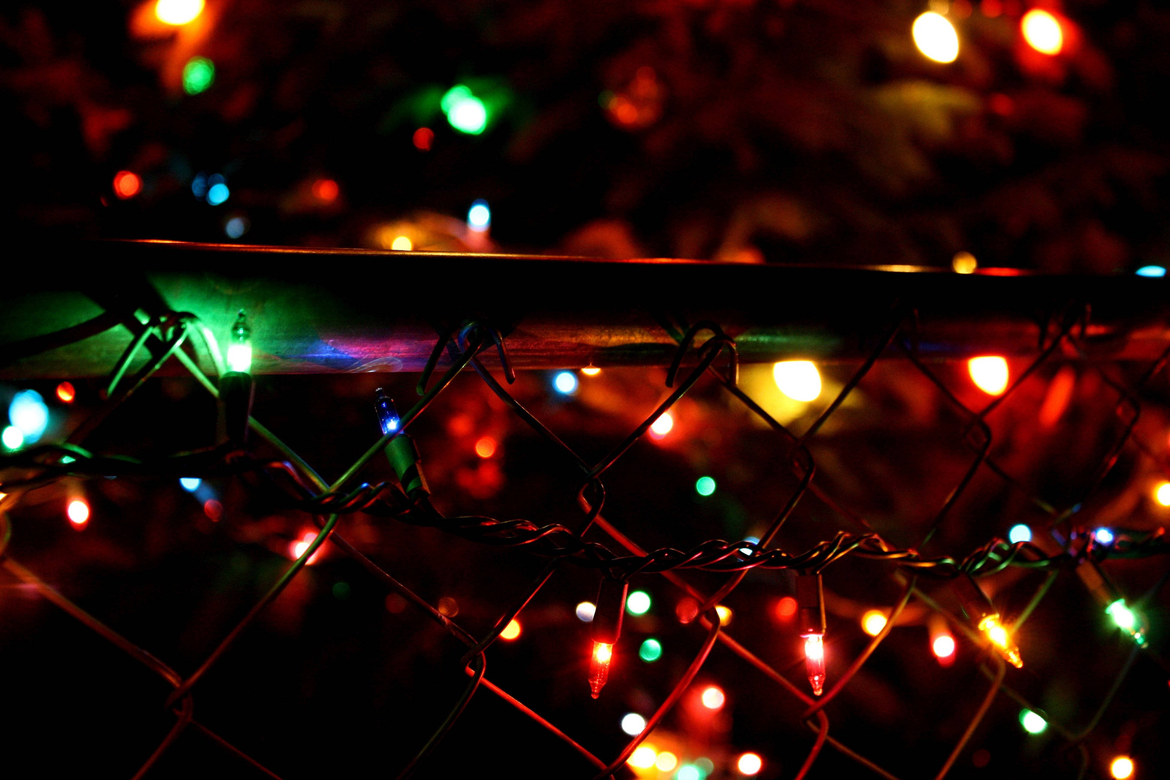 Christmas Lights Aesthetic 桌布 壁紙 — 色彩繽紛的節日燈光在夜間場景中，於深色鐵絲網圍欄後閃爍。
