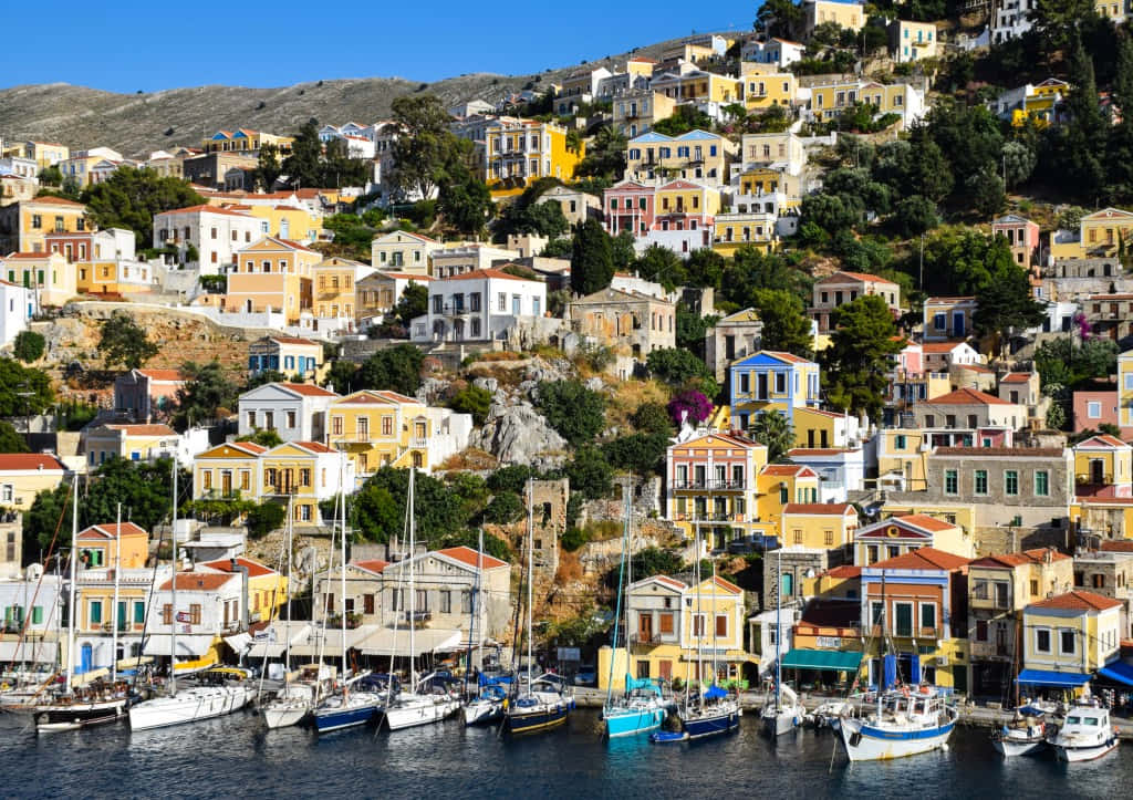 Greek Island 桌布 壁紙 — 色彩繽紛的粉彩房屋沿著陽光普照的山坡層層疊疊，俯瞰著晴朗藍天下停滿帆船的港灣。
