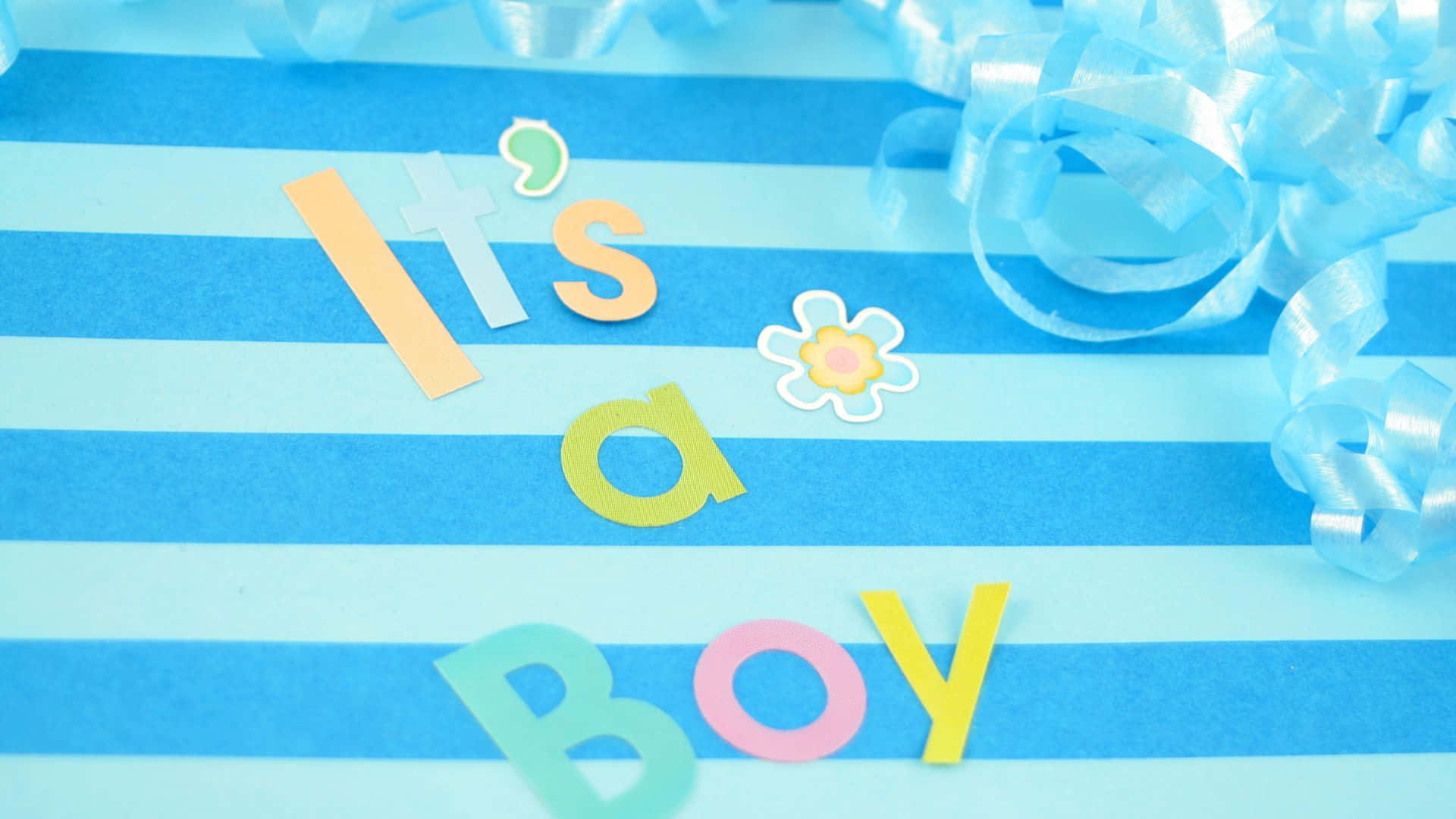 男孩 桌布 壁紙 — 色彩繽紛的紙字母拼出 'It's a Boy'，放在藍色條紋紙上，旁邊有藍色絲帶和花朵貼紙。