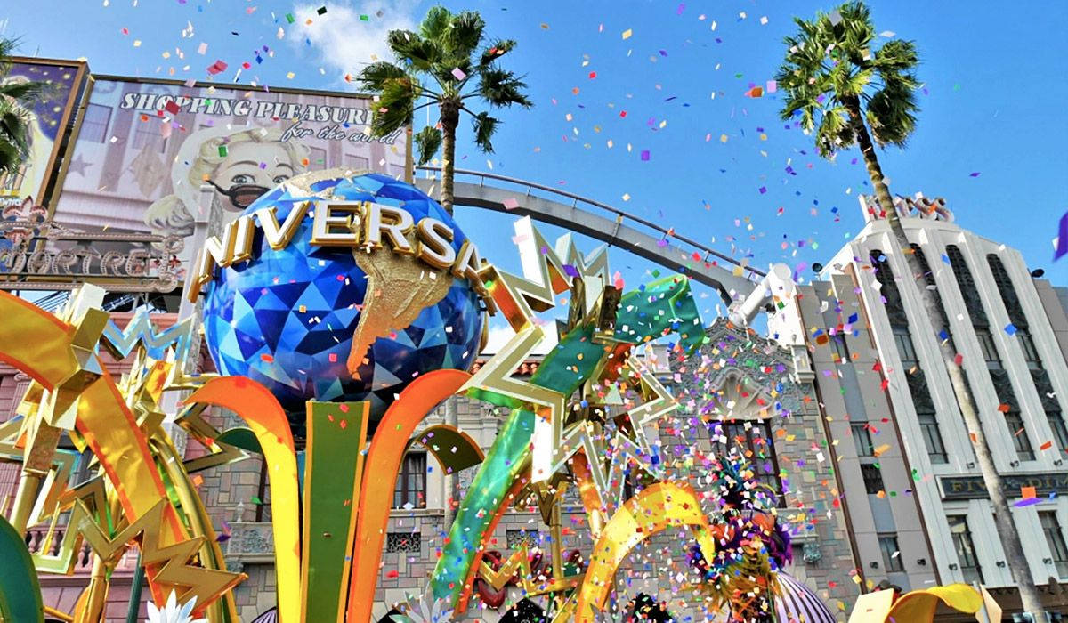 Universal 桌布 壁紙 — 色彩繽紛的紙屑在標誌性的環球影城地球儀周圍爆發，背景是藍天、棕櫚樹和主題公園建築。