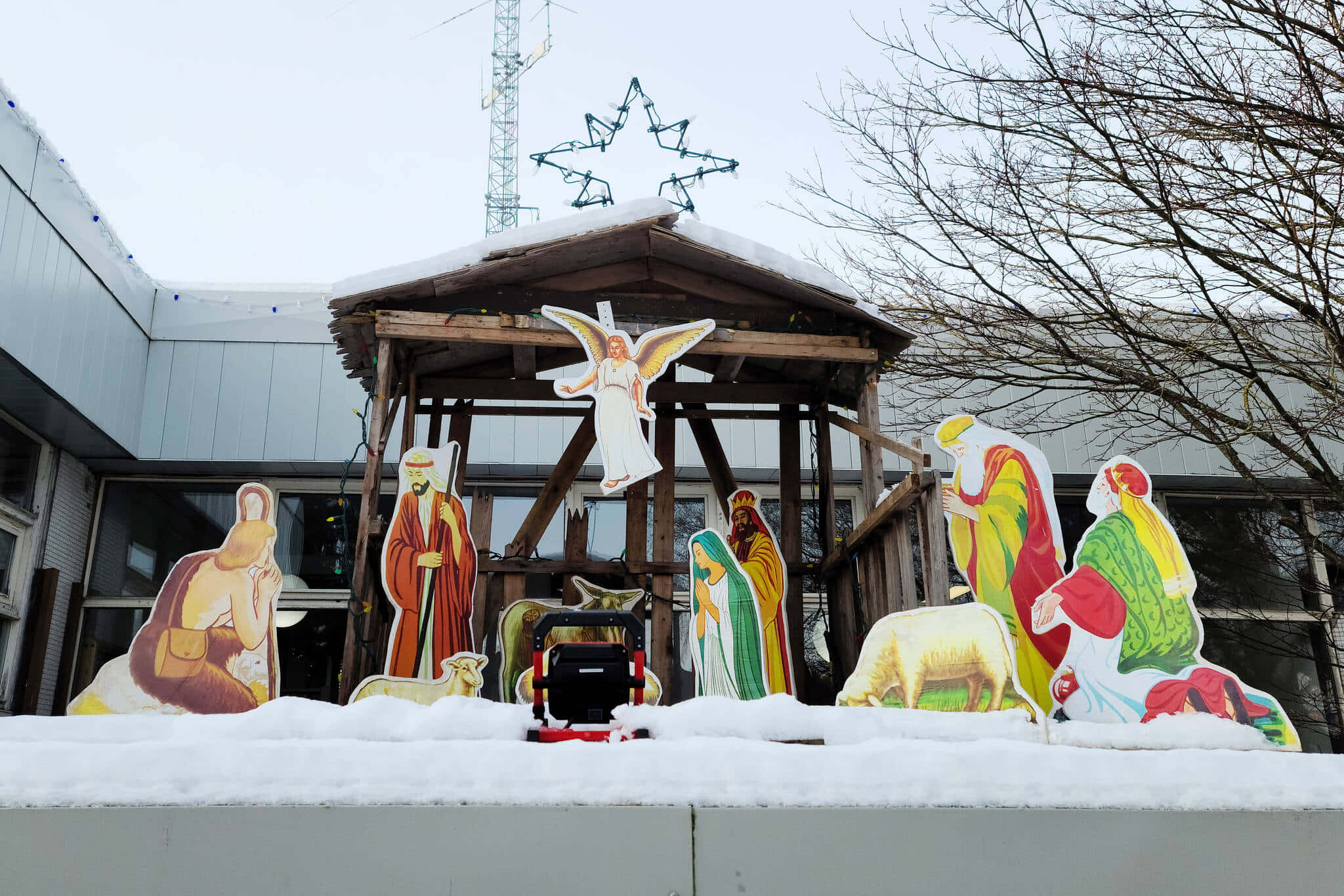 耶穌誕生場景 桌布 壁紙 — 色彩繽紛的紙板耶穌誕生人偶站在雪地上，上方有一個木製馬廄，頂部有星星，背景是光禿禿的樹木。