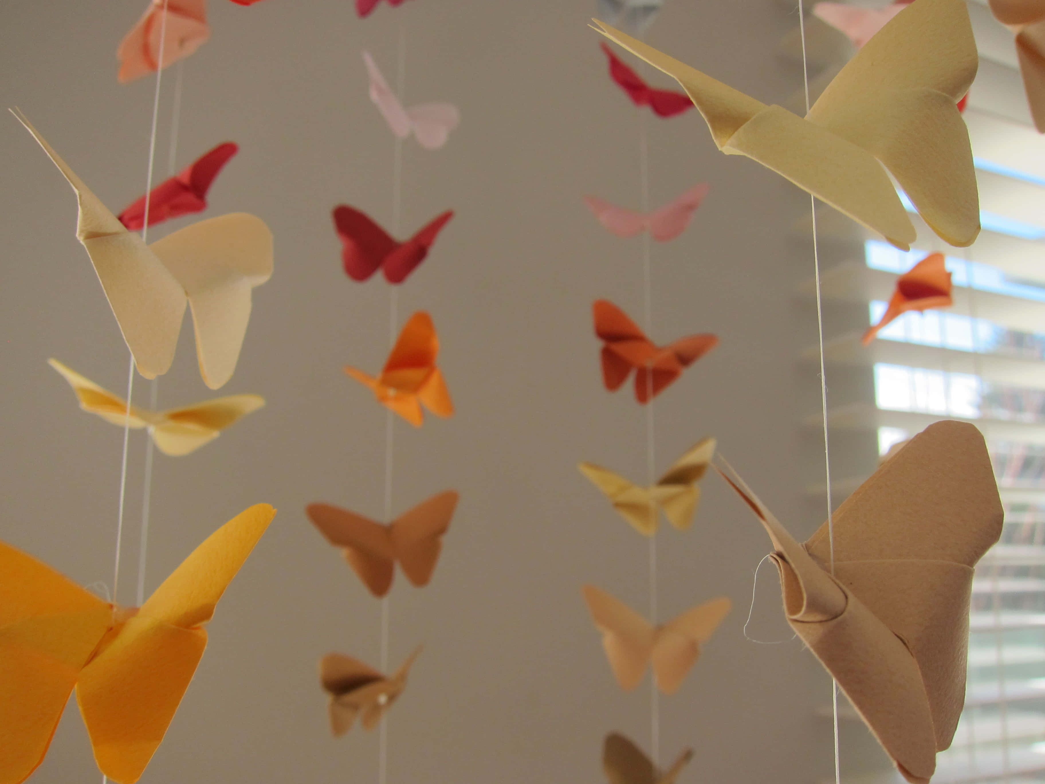 Butterfly Origami 桌布 壁紙 — 色彩繽紛的紙蝴蝶懸掛在柔和光線中，背景是帶有百葉窗的中性牆壁。
