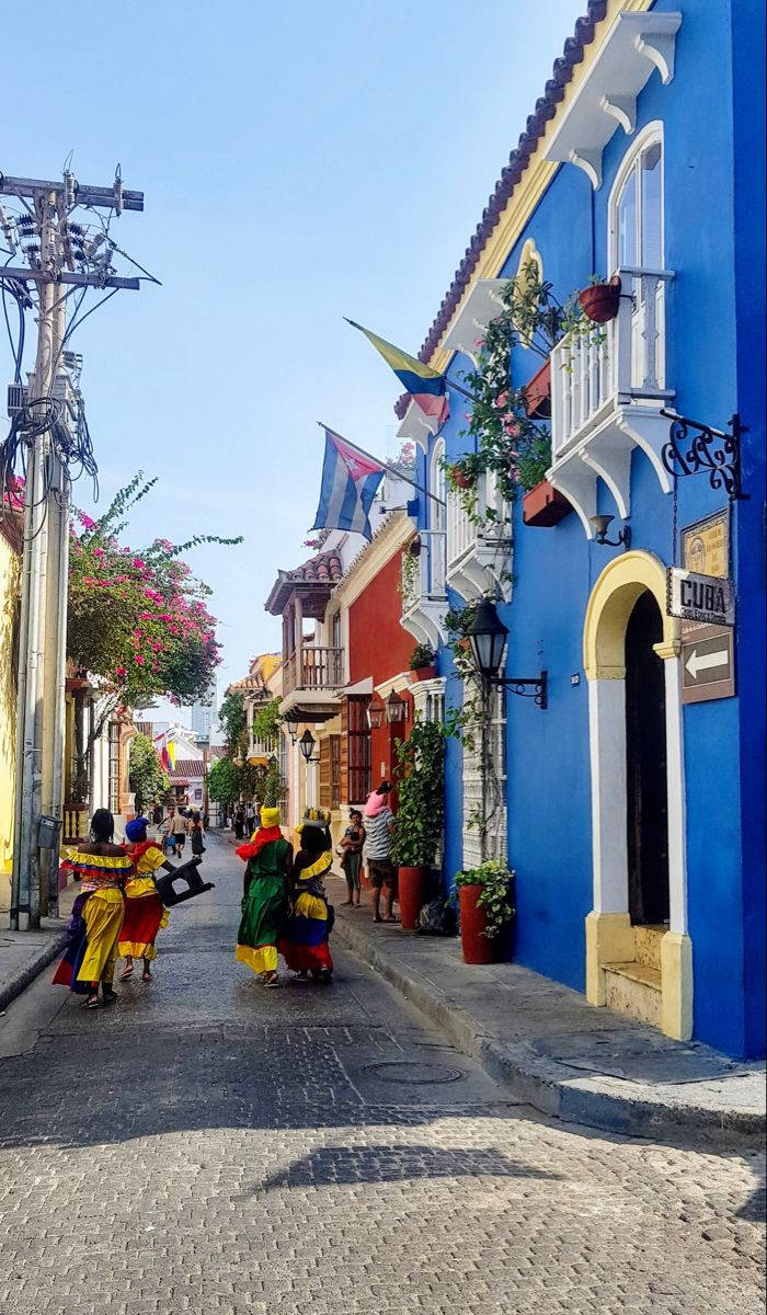 Colombia 桌布 壁紙 — 色彩繽紛的舞者穿著鮮豔服裝，在晴朗的藍天下，沿著一條鋪滿鵝卵石的街道遊行，街道兩旁是明亮的藍色和紅色殖民時期建築。