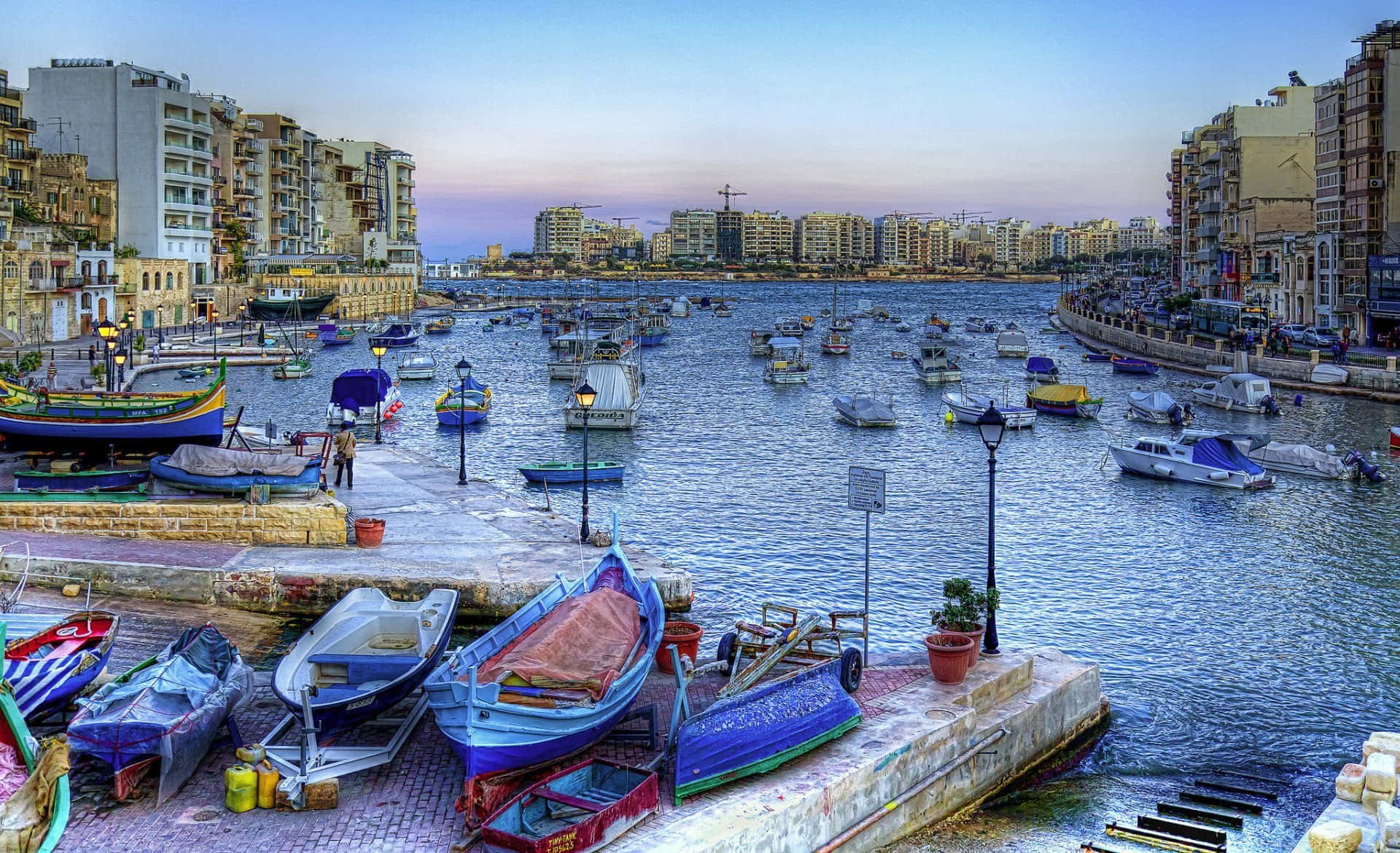 Malta 桌布 壁紙 — 色彩繽紛的船隻點綴著黃昏時分繁忙的港口，海濱兩旁是城市建築。