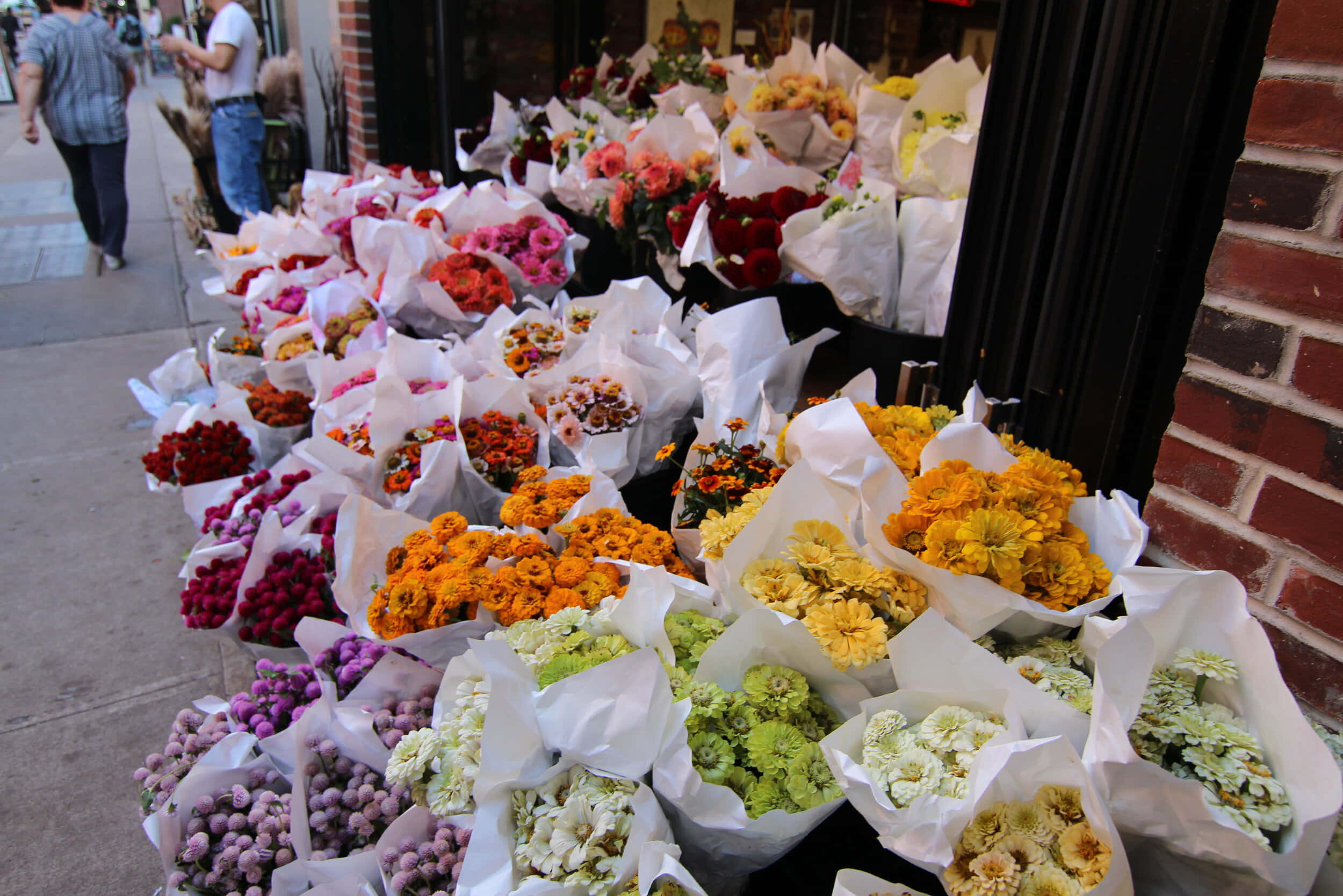 Flower Market 桌布 壁紙 — 色彩繽紛的花束用白紙包著，排列在繁忙城市人行道的磚牆店鋪前面。