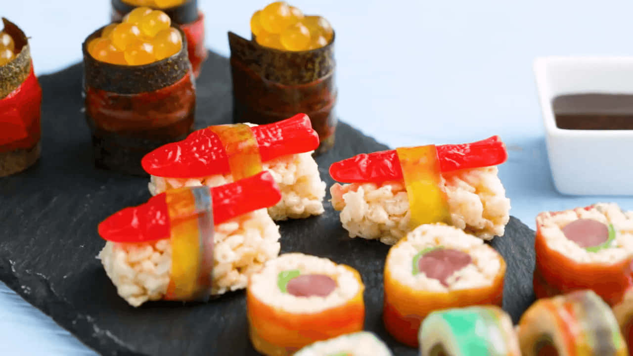 Sushi 桌布 壁紙 — 色彩繽紛的軟糖壽司捲和魚子醬捲，排列在黑色石板上，配以醬油。