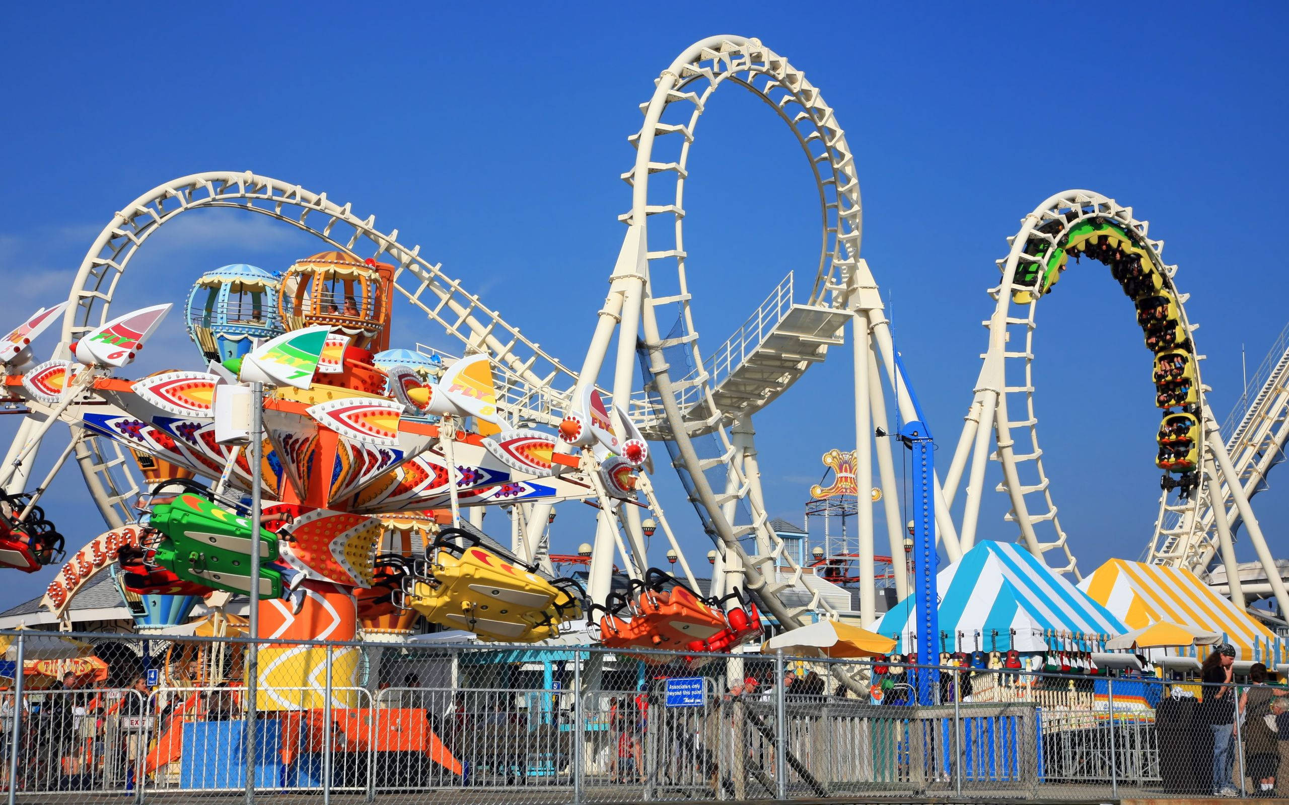 Roller Coaster 桌布 壁紙 — 色彩繽紛的遊樂園遊樂設施在晴朗的藍天下蜿蜒轉動，伴隨著多彩的景點和行駛中的雲霄飛車。