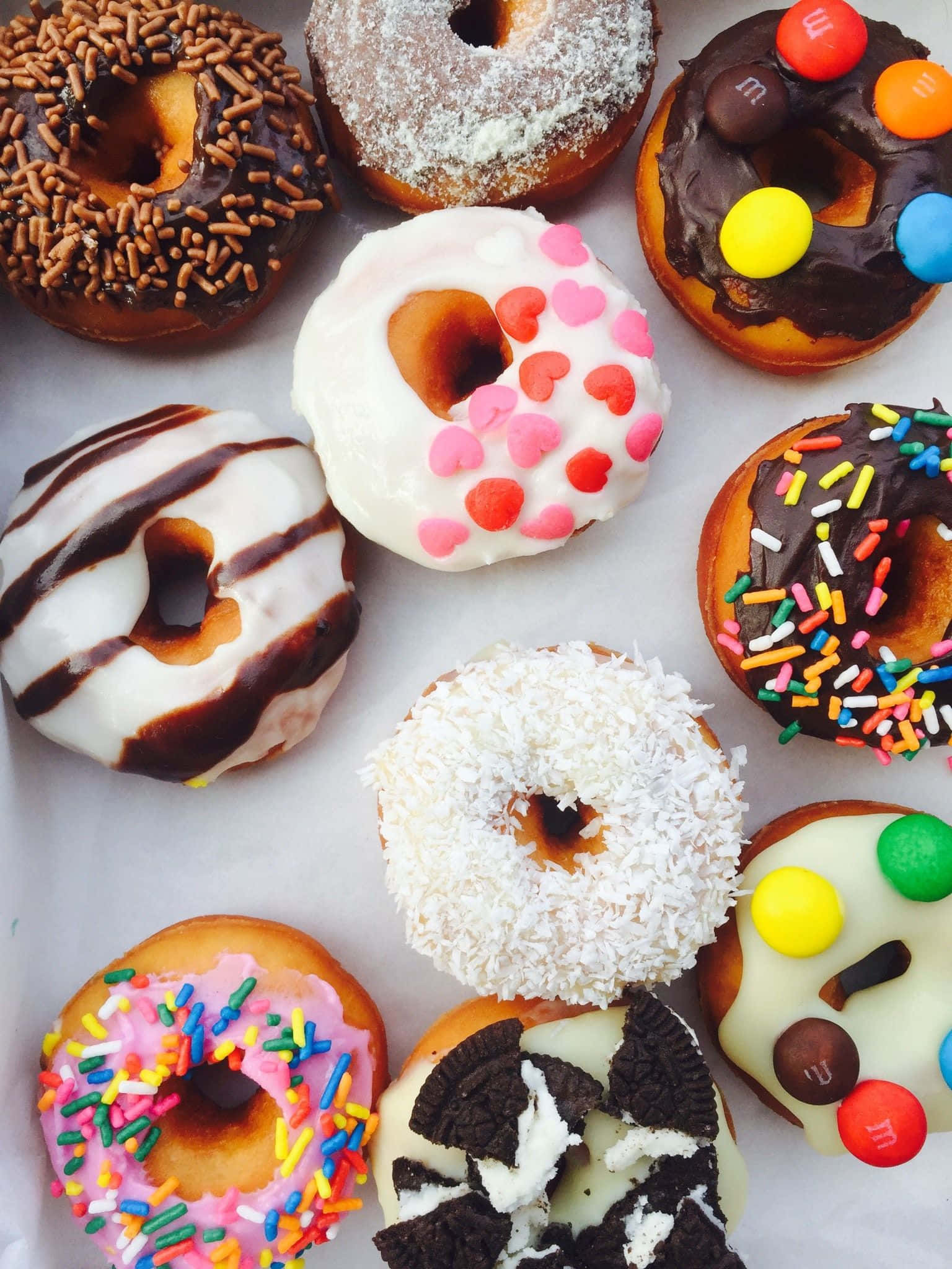 donuts 桌布 壁紙 — 色彩繽紛的釉面及裝飾甜甜圈，配上糖針、糖果和巧克力配料，整齊排列在白色烘焙紙上。