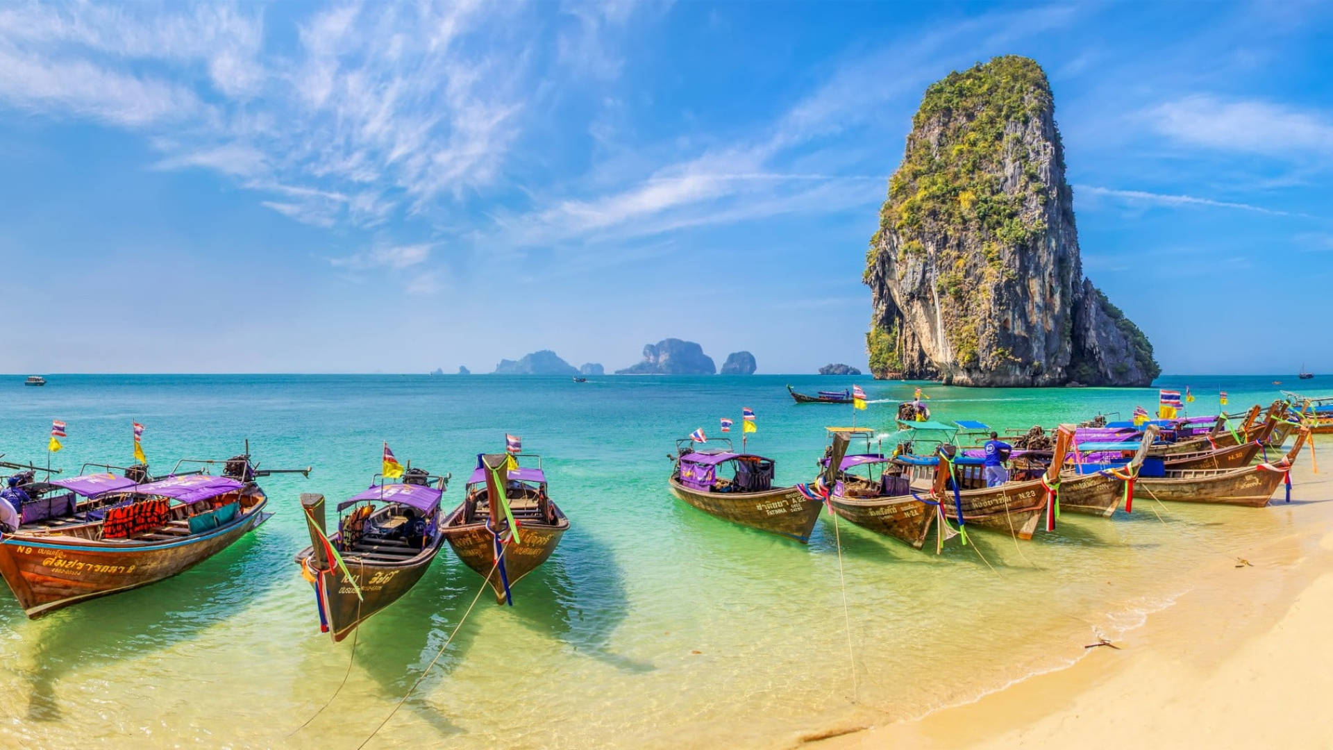Thailand 桌布 壁紙 — 色彩繽紛的長尾船停泊在淺藍綠色的海水中，旁邊是金色沙灘和 towering 石灰岩懸崖，在明亮的藍天下。