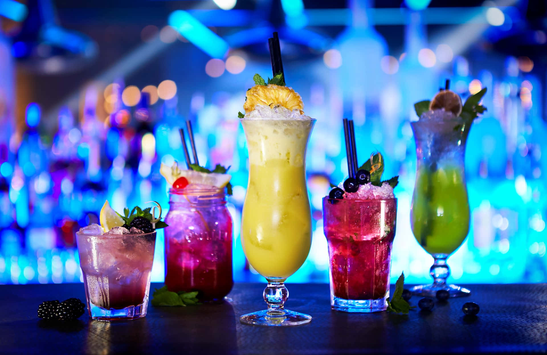 drinks 桌布 壁紙 — 色彩繽紛的雞尾酒盛載於各式玻璃杯中，飾以水果和薄荷，放在吧檯上，背景是藍色燈光下模糊的酒瓶。