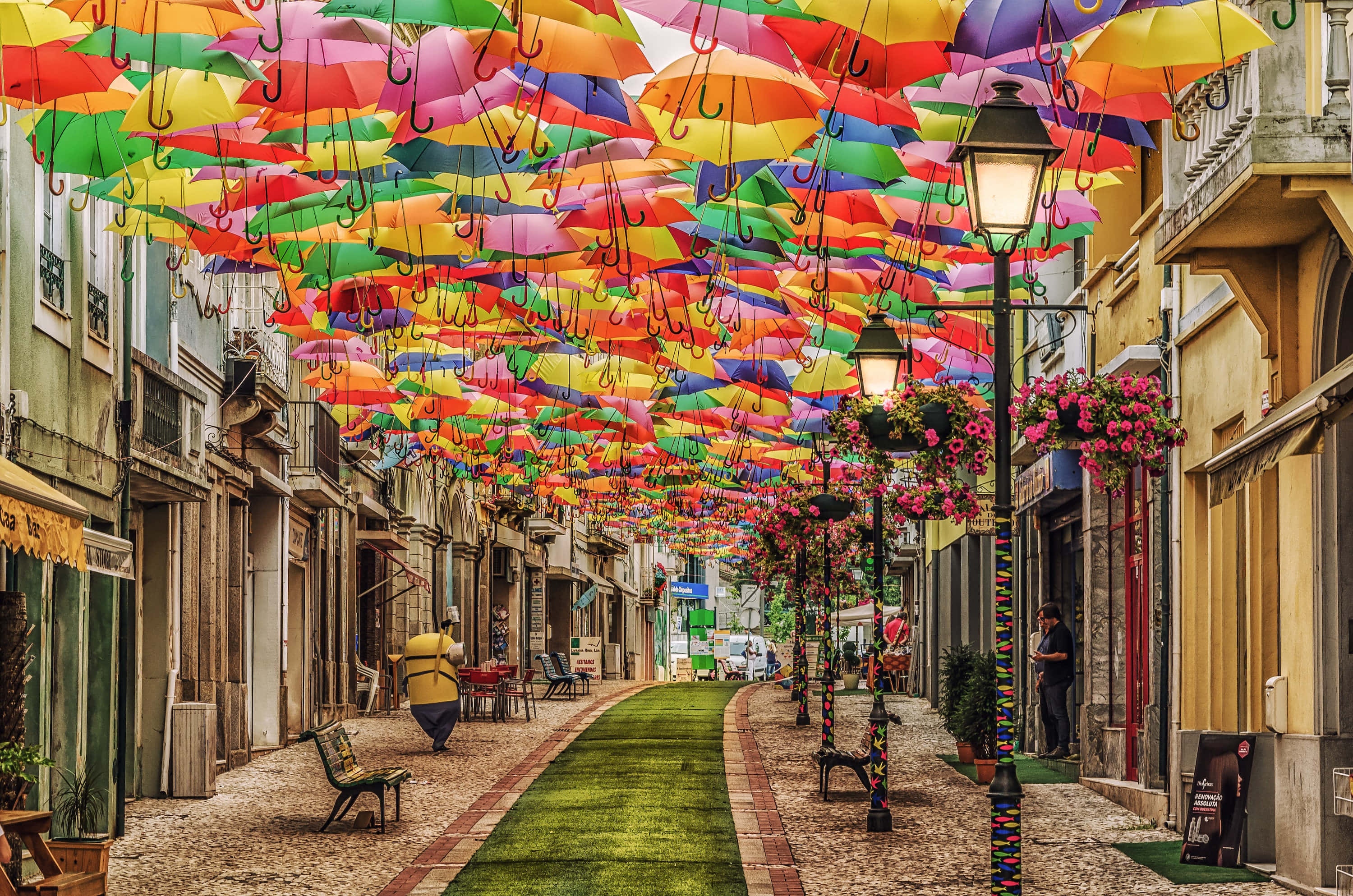 Umbrella 桌布 壁紙 — 色彩繽紛的雨傘形成充滿活力的街道天篷，拱形覆蓋在鵝卵石小巷上，掛有花朵和溫暖的燈光。