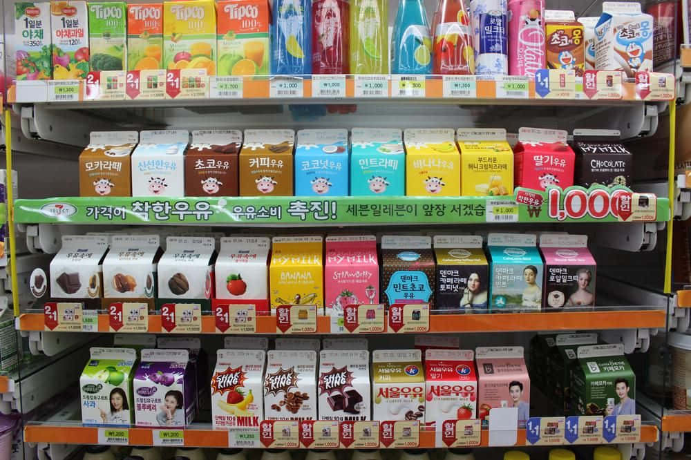 7-ELEVEN 桌布 壁紙 — 色彩繽紛的韓國飲料和牛奶盒整齊地排列在超市貨架上，附有價格標籤和促銷橫幅。