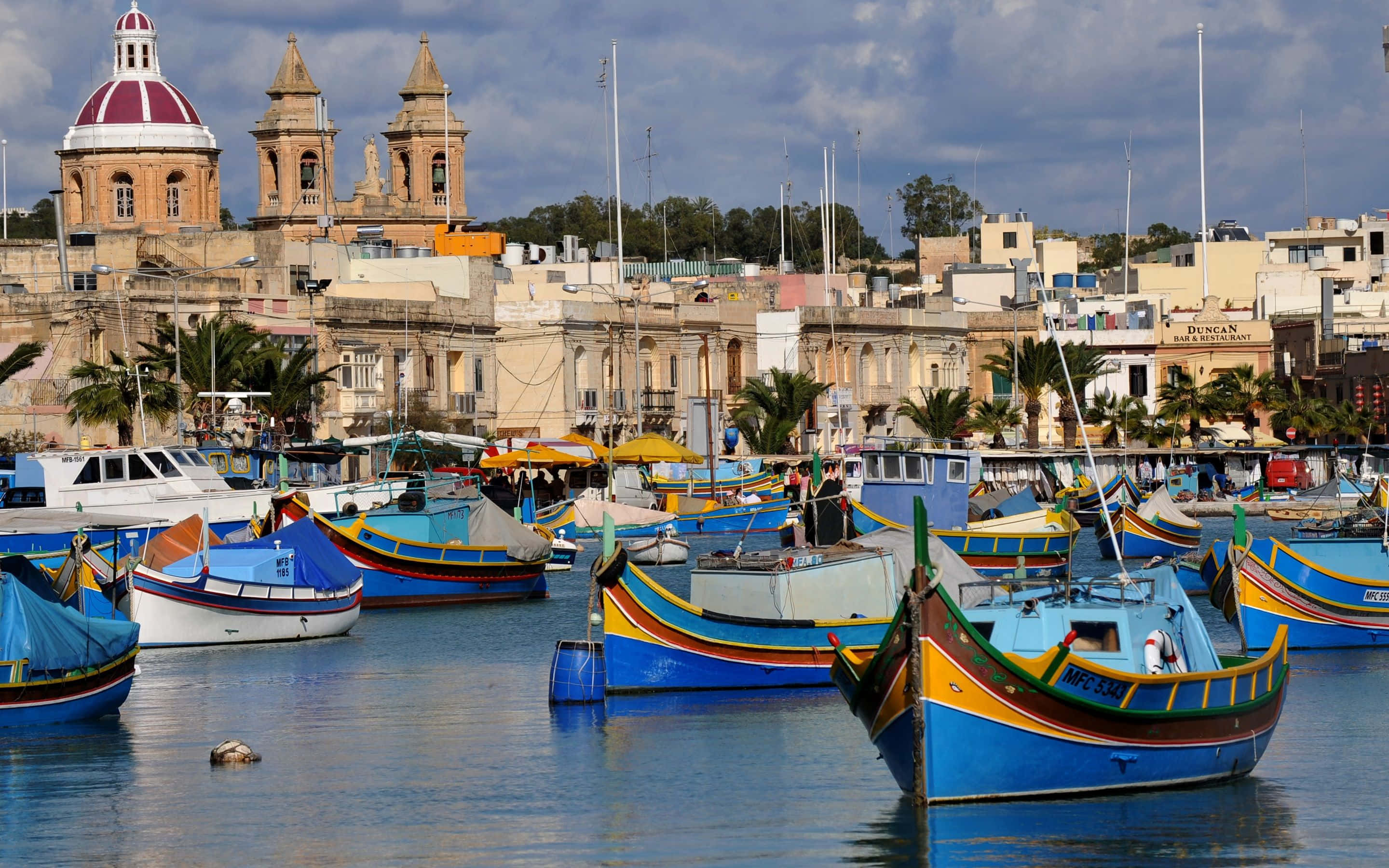 Malta 桌布 壁紙 — 色彩鮮豔的藍黃色馬耳他魯祖船隻在繁忙的港口中漂浮，背景是歷史悠久的圓頂教堂和陽光普照的建築物。