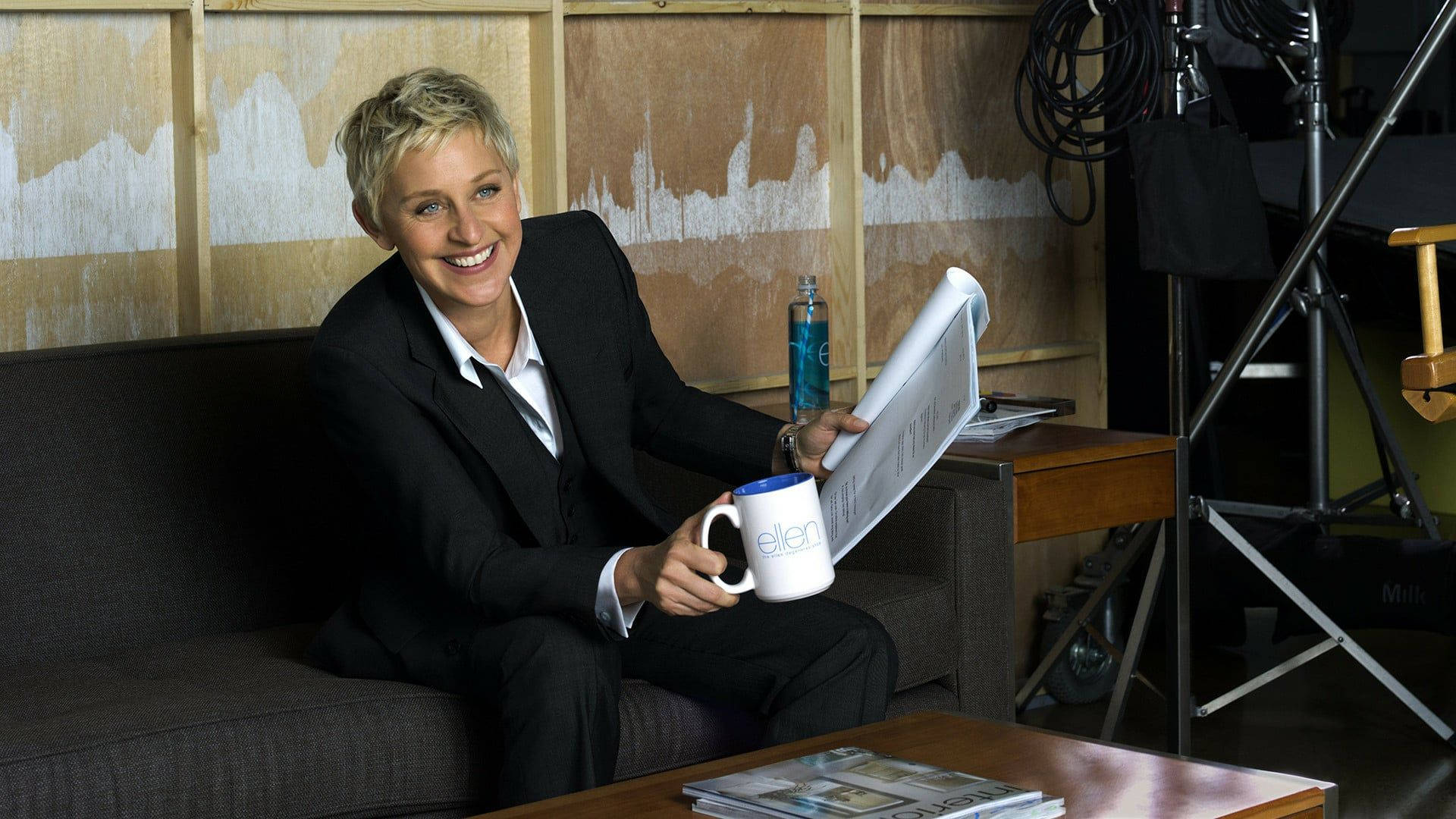 Ellen Degeneres 桌布 壁紙 — 艾倫·狄珍妮在攝影棚的沙發上拿著馬克杯和報紙，露出溫暖的笑容。